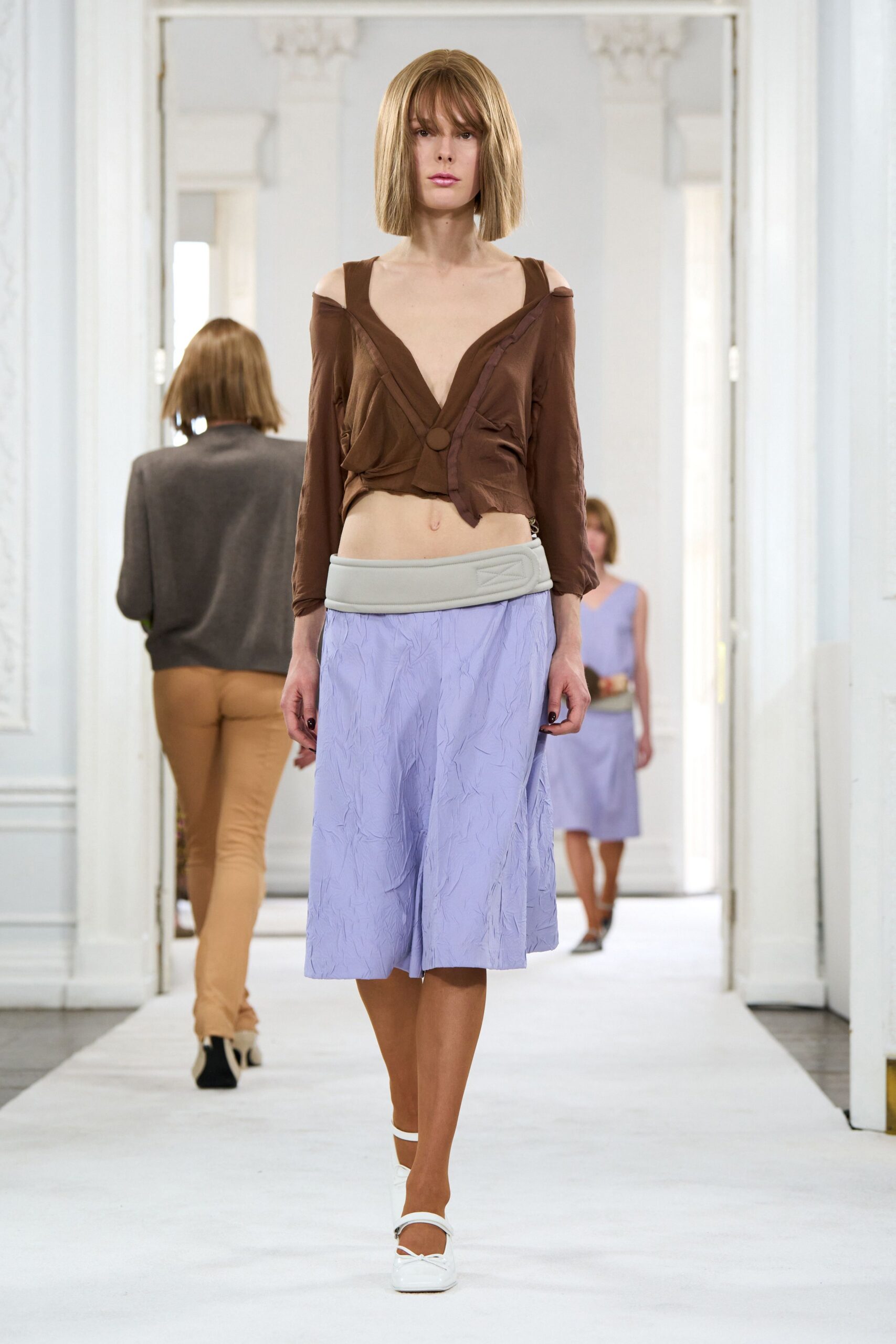 Chocolate off-shoulder top in slinky jersey, twisted to a single covered button. Pale lilac crinkled skirt with a padded dove-grey waistband. White ankle-strap flats, squared toe. Morning after the hotel wedding, carrying your shoes by noon.