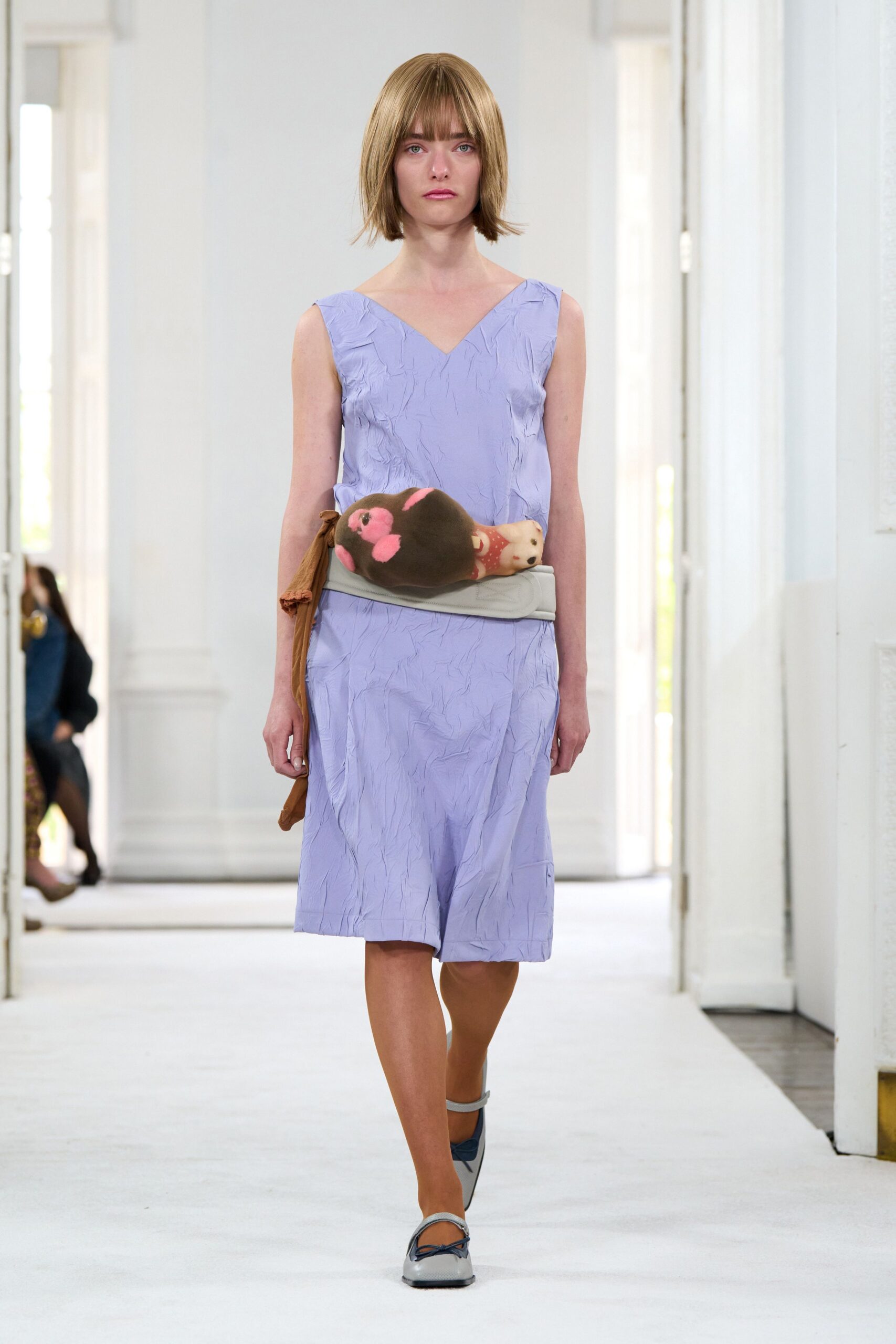Lavender V-neck dress in crinkled fabric, hanging loose to the knee. Pale padded waist belt clasping a plush mole and tiny teddy. Grey ankle-strap flats with a dark bow across the vamp. For crossing a museum corridor with a toy tucked under your arm.