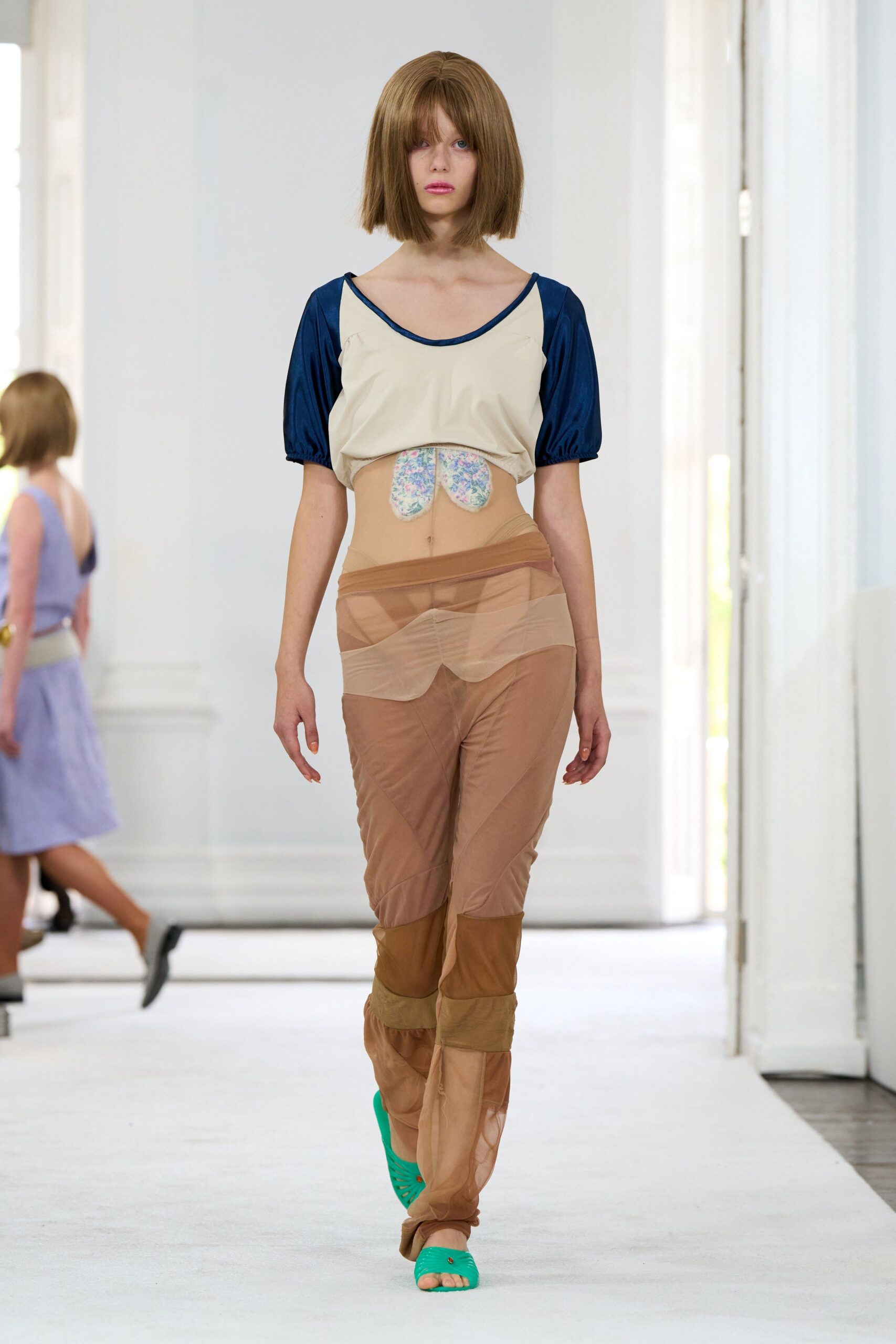 Cream jersey top with cobalt satin puff sleeves and a dipped hem. Two floral padded patches hanging at the bare midriff. Layered nude mesh trousers, sheer bands crossing low over the hips. Bright green jelly sandals with a tiny rivet at the strap. Walking back from the lido with sunscreen drying sticky on your skin.