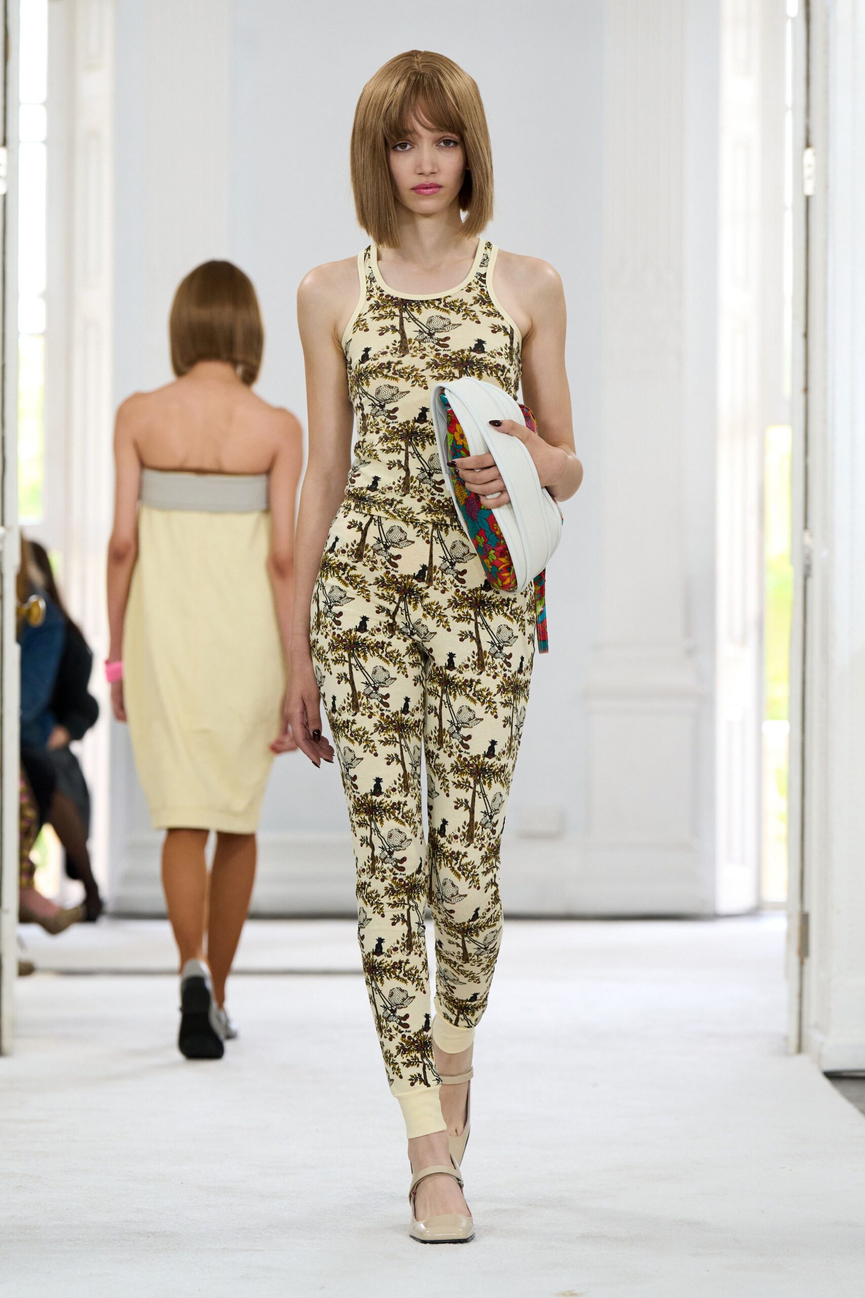 Cream stretch jersey jumpsuit, racerback cut with brown bird-and-vine print. Quilted white crescent bag, floral lining flashing at the zipper edge. Cream patent Mary Janes with a narrow instep strap. Walking the long hallway to a conservatory lunch after rain.