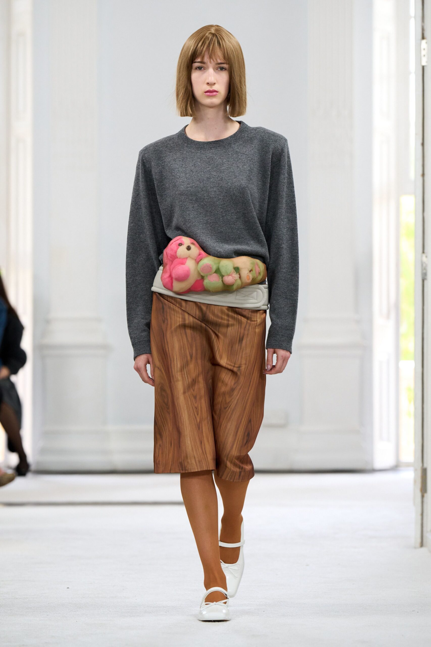 Charcoal fine-knit sweater, slightly dropped at the shoulder. Pink stuffed dog strapped over a padded white belt. Brown knee-length skirt printed with swirling woodgrain. Opaque caramel tights. White patent Mary Janes with a tiny bow. For walking into a rental cabin whose walls are all knotty pine.