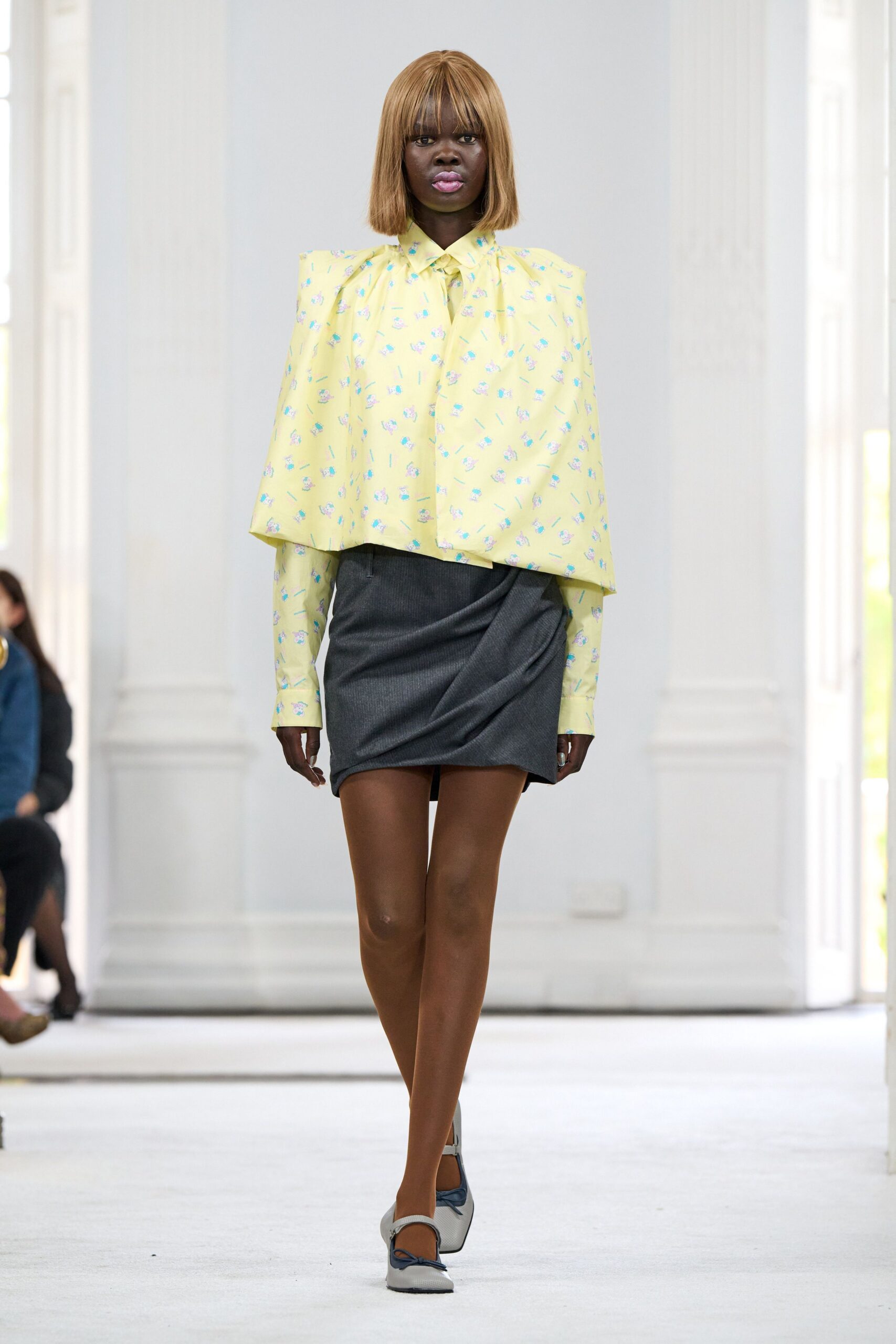 Butter-yellow blouse printed with tiny blue bouquets, cape panel skimming past the elbows. Charcoal pinstriped mini, draped into a diagonal fold across the hips. Grey leather mary janes with navy bow trim at the toe. Walking into a spring office party still carrying your coat check ticket.