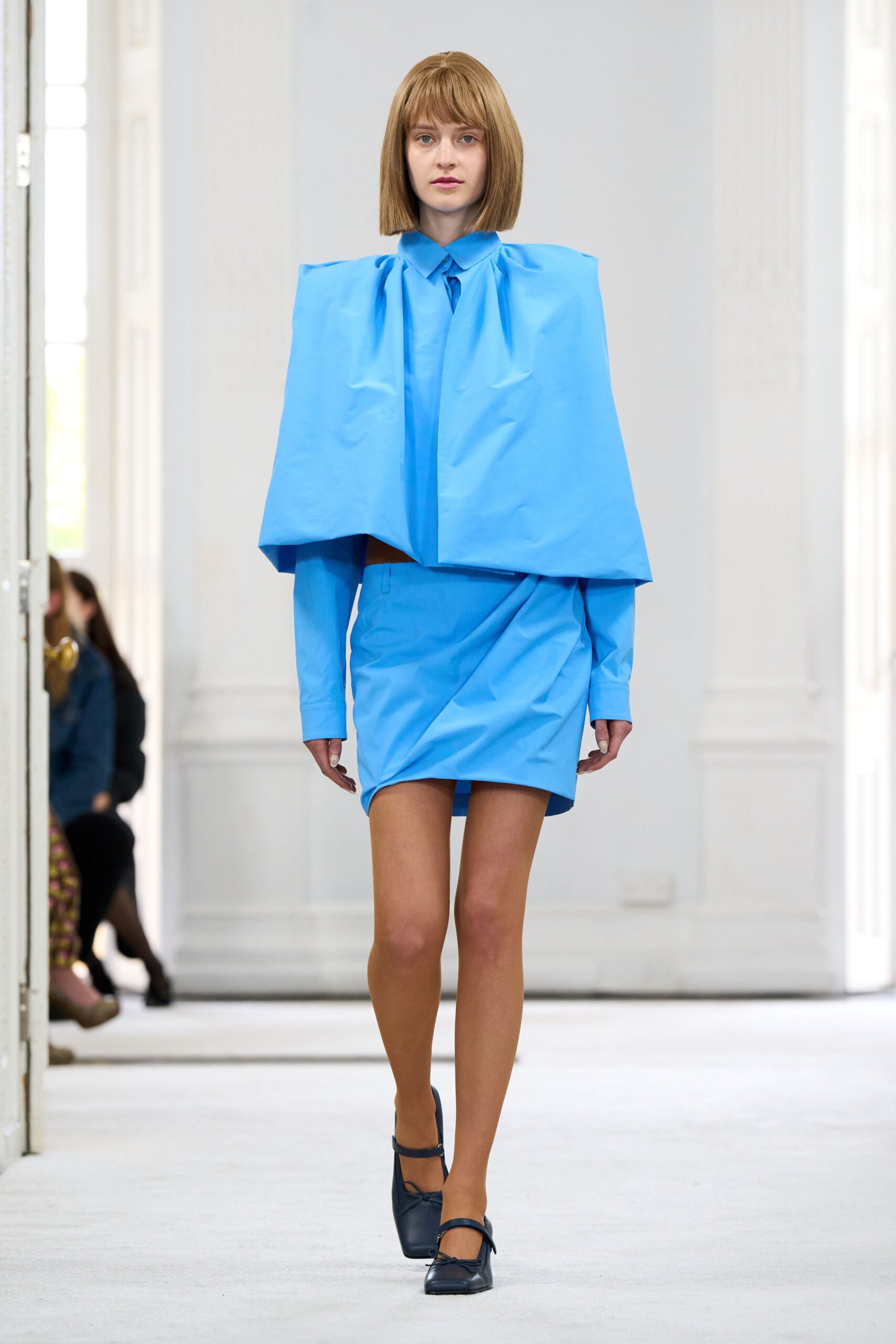 Sky-blue cotton poplin blown into a boxy cape over a pointed collar. Matching wrap mini, diagonally ruched with a narrow strip hanging below the hem. Black leather Mary Janes with squared toes and tiny bow ties. Crossing a marble atrium before the offices open and the echo dies down.