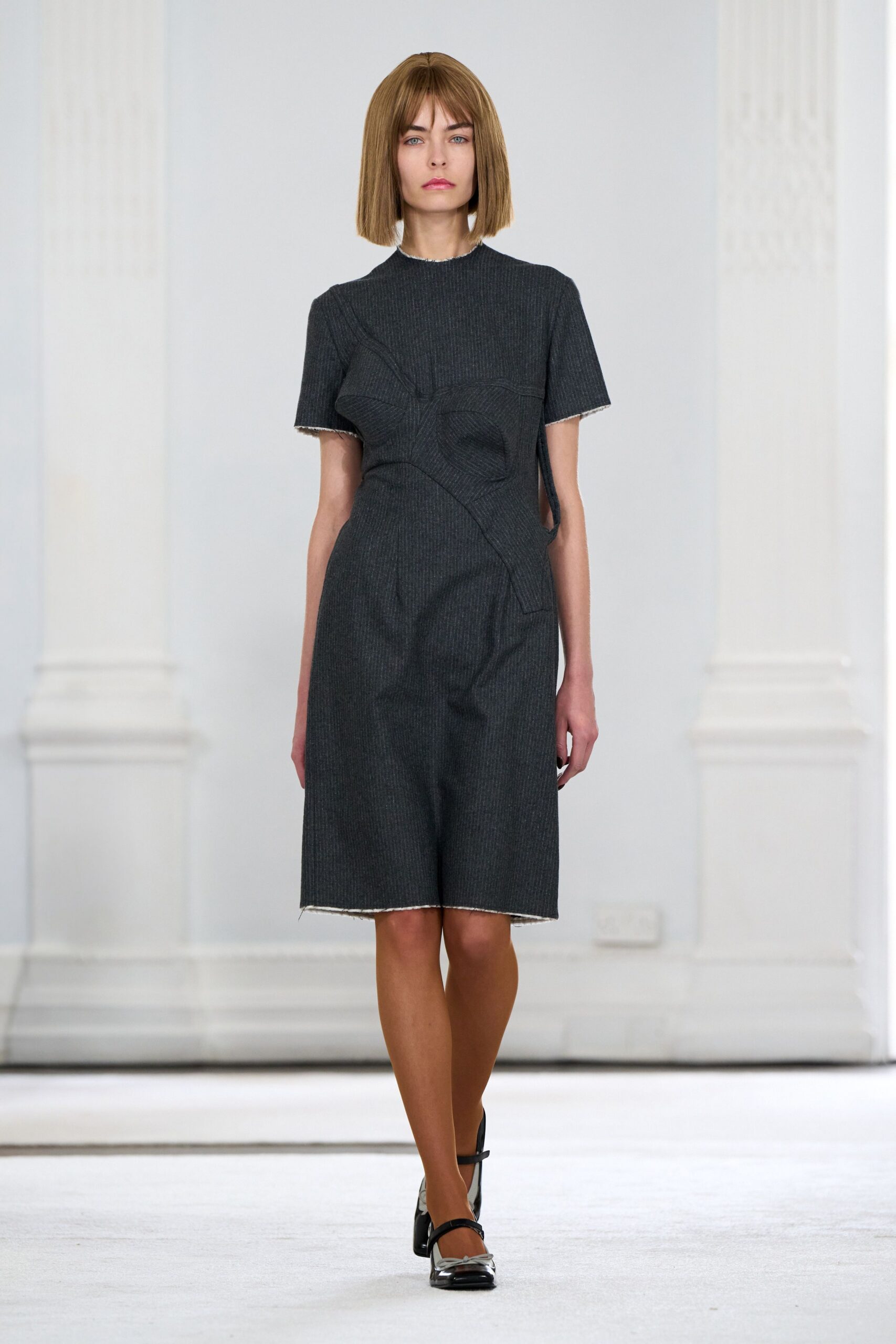 Charcoal pinstriped dress with sculpted bust seams and raw ivory edges. Black patent Mary Janes with a square, silvered toe cap. Walking into a municipal chamber just after the radiators kick on.