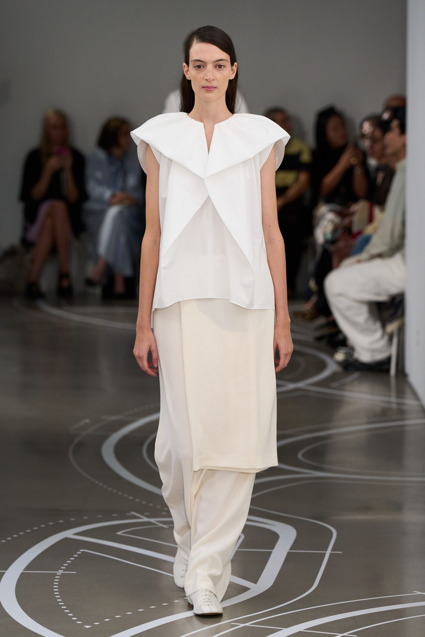 White sleeveless top with a giant folded collar crossing into sharp points. Ivory column skirt in fluid crepe, hanging straight over the hips. Wide white trousers pooling softly below the skirt hem. Square-toe white lace-ups, almost hidden under the break. Museum atrium at opening hour, footsteps echoing before anyone speaks.