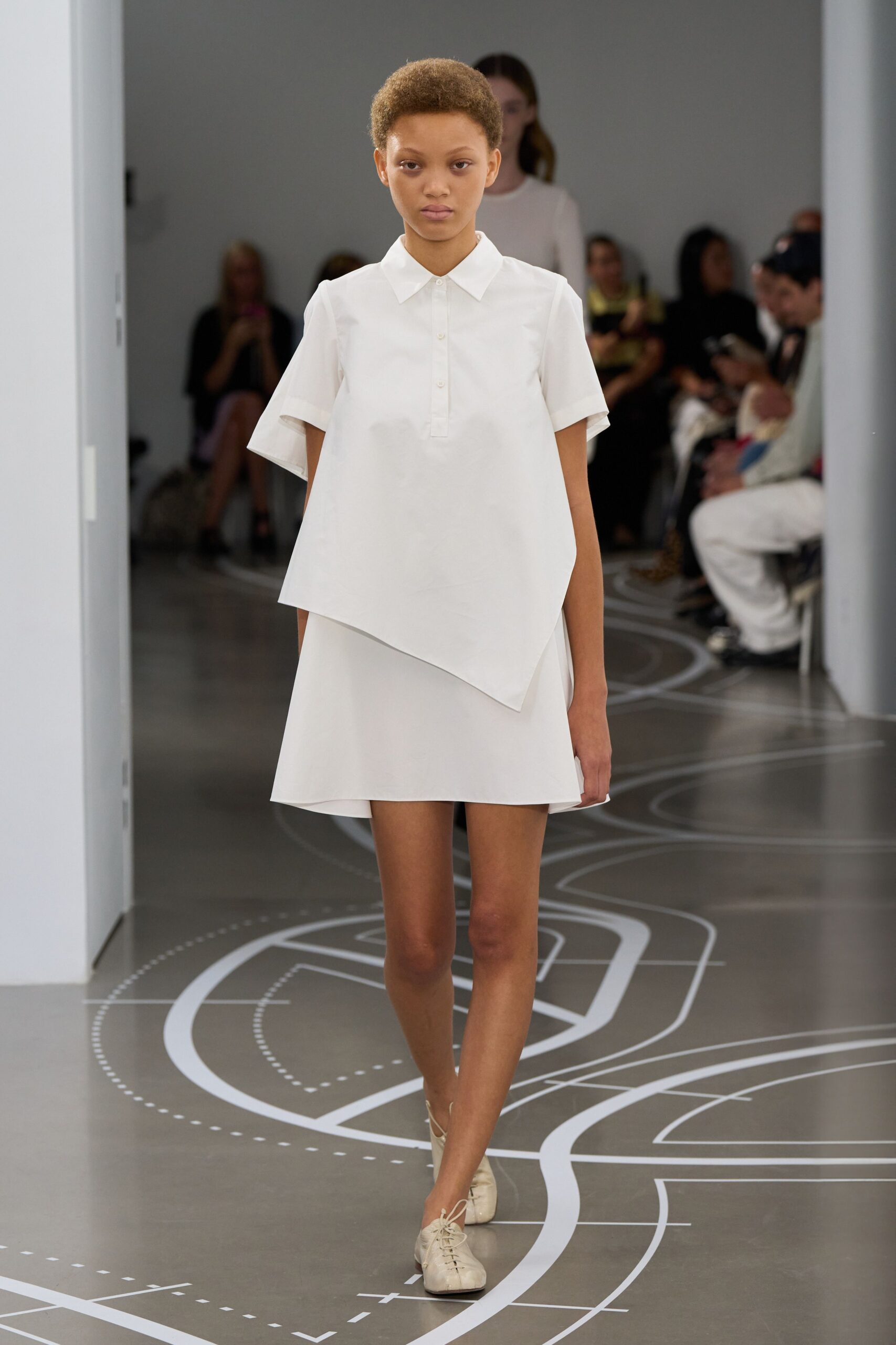 White cotton popover with a pointed collar and angled overlapping hem. Matching A-line mini, cleanly cut with a slight swing. Cream leather lace-ups, softly creased across the vamp. Walking through a museum atrium before the school groups arrive.
