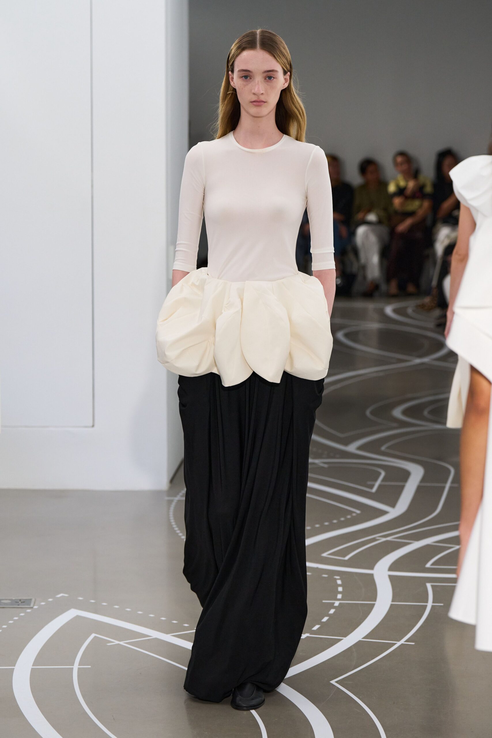 Sheer ivory jersey top with close crew neck and elbow sleeves. Quilted cream hip peplum stuffed into rounded petal-like poufs. Black floor-length skirt falling in heavy off-center jersey folds. Black square-toe flats peeking from the hem. Crossing a museum atrium while the echo gets there first.