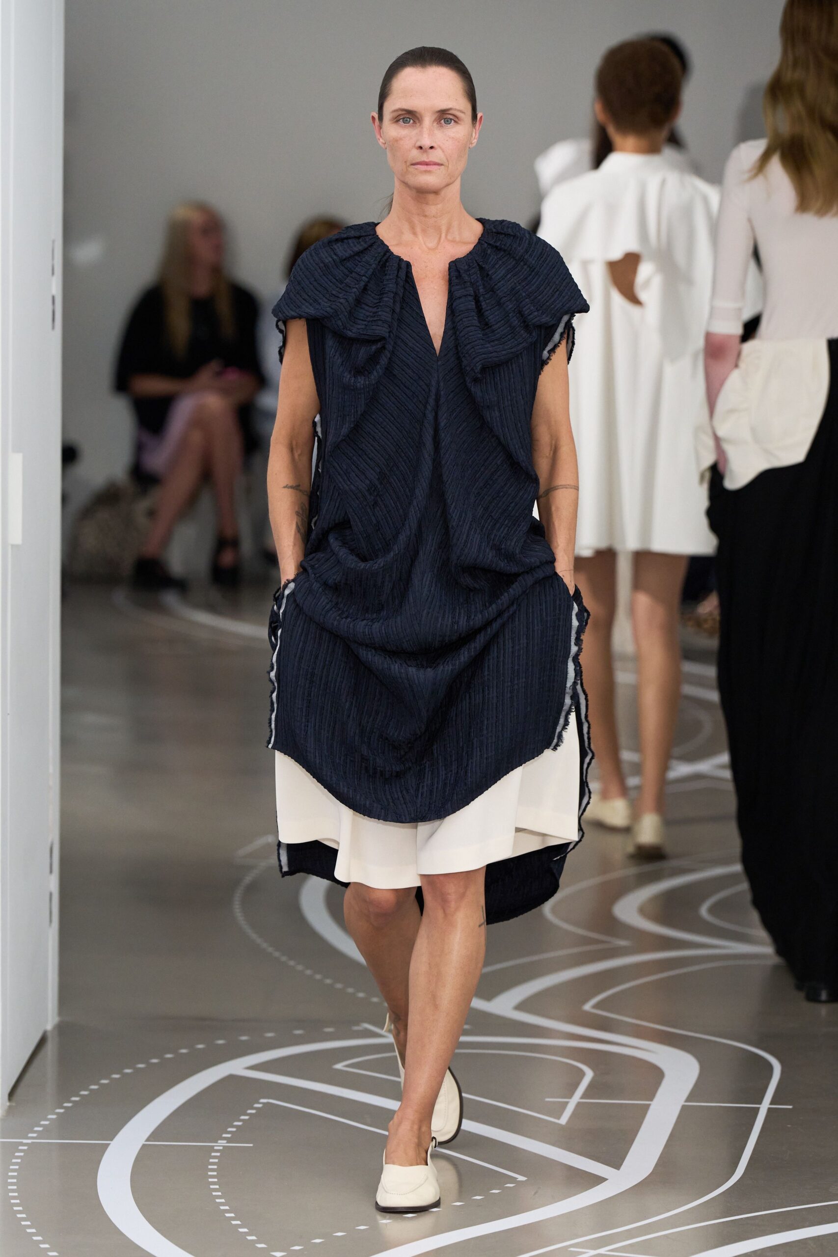 Ink-black crinkled tunic with a ruched cape collar and frayed edges. Chalk tailored shorts peeking below the uneven hem. Cream loafers with squared vamps and dark soles. Crossing a museum atrium before the air conditioning fully wakes up.