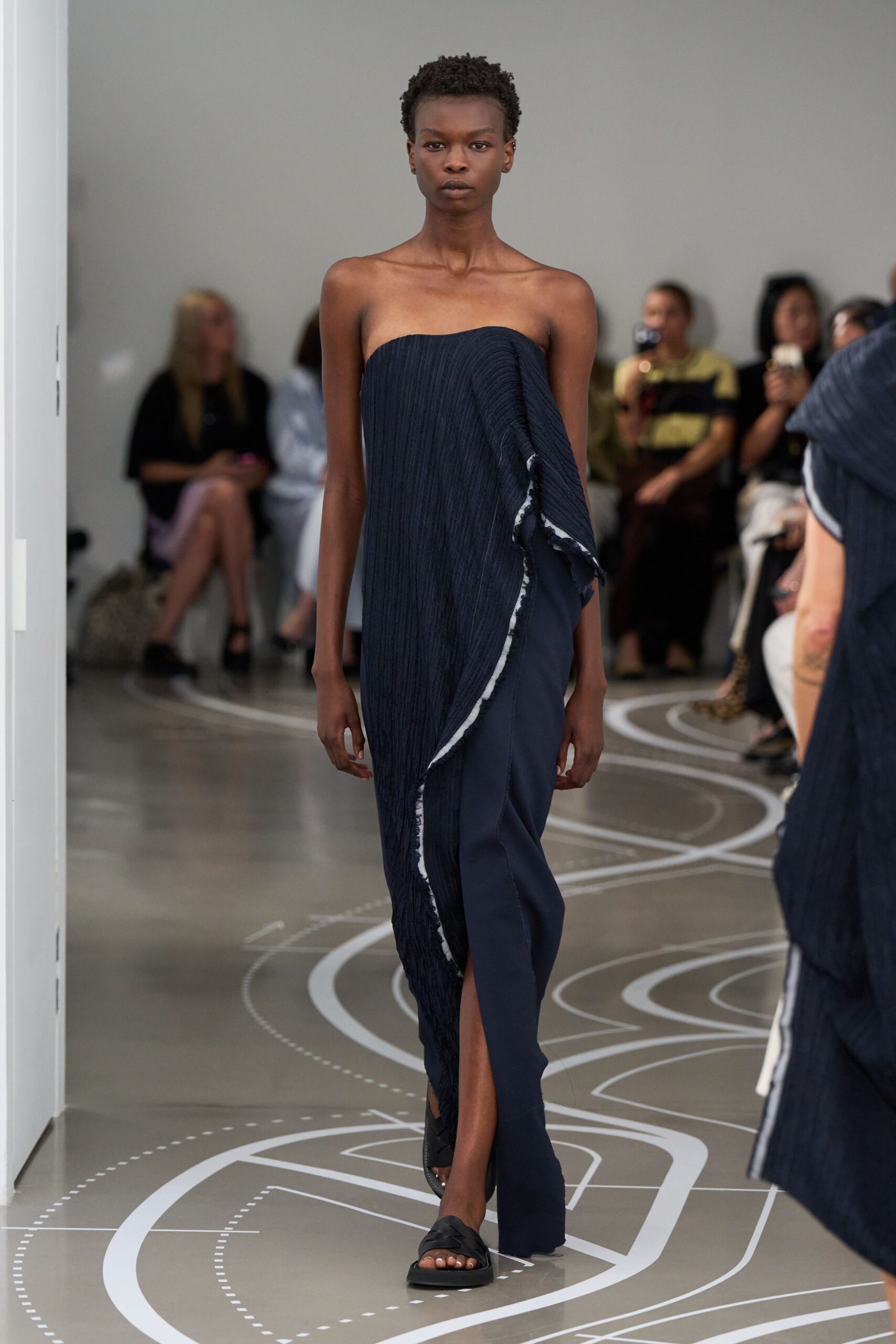 Strapless navy column dress with a crinkled overlay curling off one side. Black padded slides, square at the toe. Walking the hotel corridor barefoot later, duvet still creased on your skin.