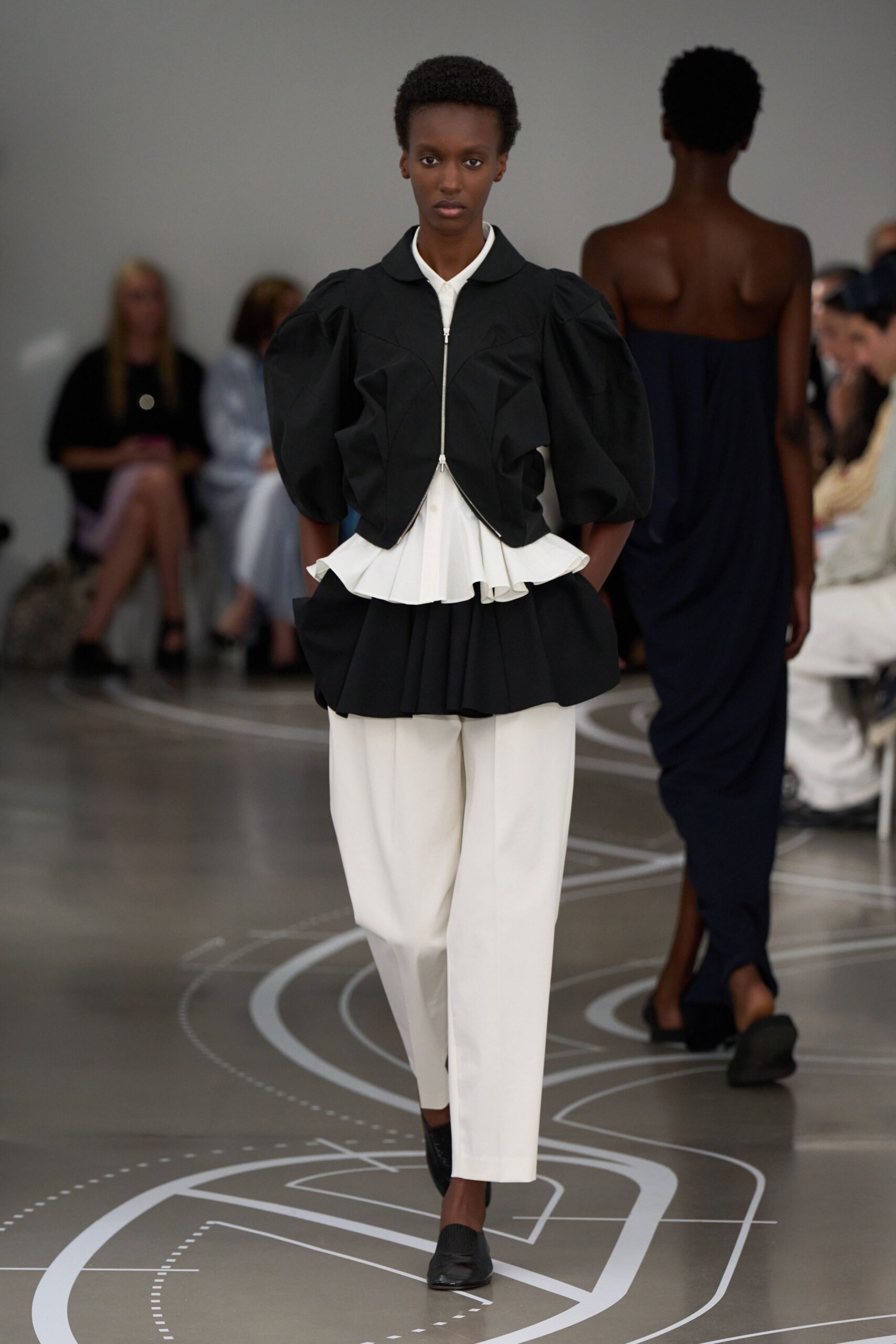 Black cropped jacket with carved puff sleeves and a silver zip. White pointed-collar shirt ending in a pleated peplum flare. Black peplum overskirt jutting out in stiff folded volume. Cream wide trousers with a clean break above the foot. Black knit flats. Crossing a museum atrium before the rain reaches the glass roof.