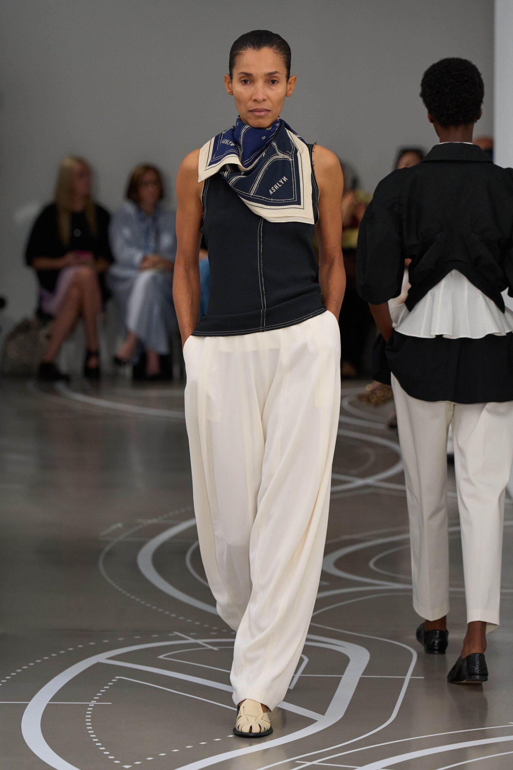 Navy silk square scarf, map-like white border folded at the throat. Ink sleeveless shell with contrast topstitching down the center seam. Ivory fluid trousers, cut loose through the leg with hands-in pockets. Cream woven leather flats with black soles. Walking a long ferry corridor before the island comes into view.