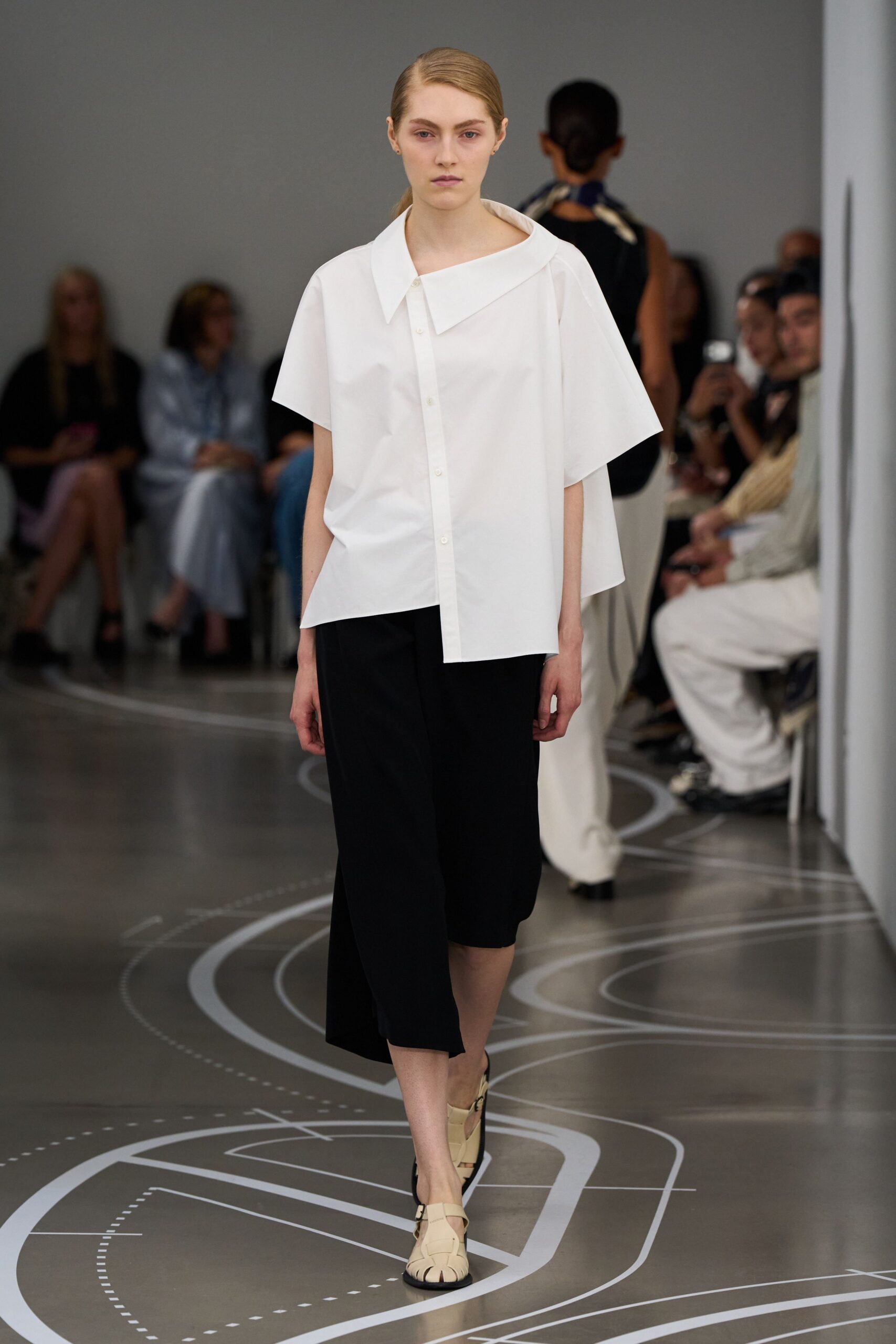 Crisp white poplin shirt with an oversized skewed collar and cape back. Black mid-calf culottes, cut wide and flat through the front. Cream fisherman sandals with thick woven straps and black soles. Crossing a quiet museum atrium before the school groups arrive.