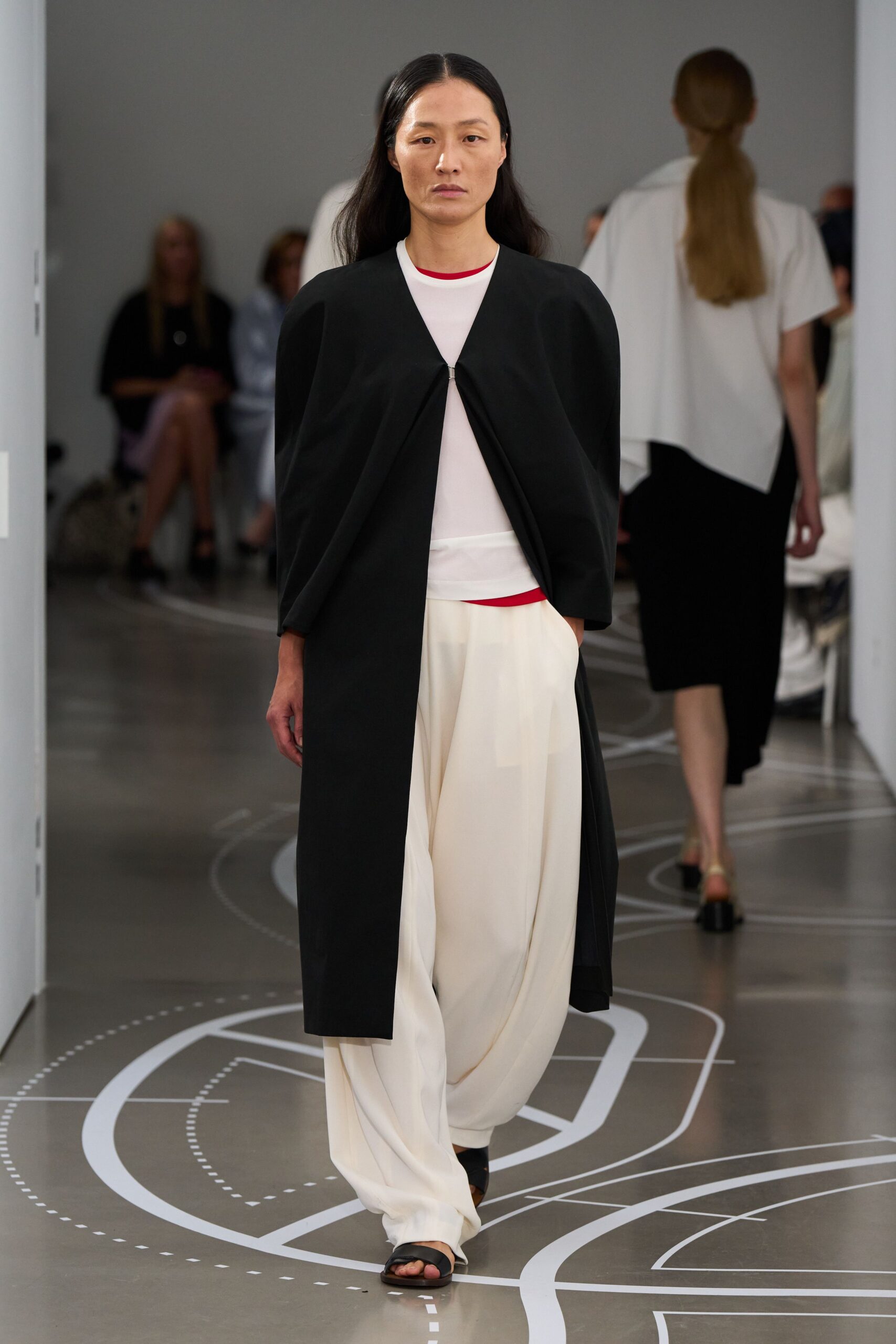 Black collarless coat, pinched shut at the chest with a tiny metal clasp. Sheer white top with a red bound neckline and matching hem peeking out. Cream fluid trousers, dropped into soft folds that puddle over the foot. Black slide sandals. Walking museum corridors before opening, floor plan still taped underfoot.