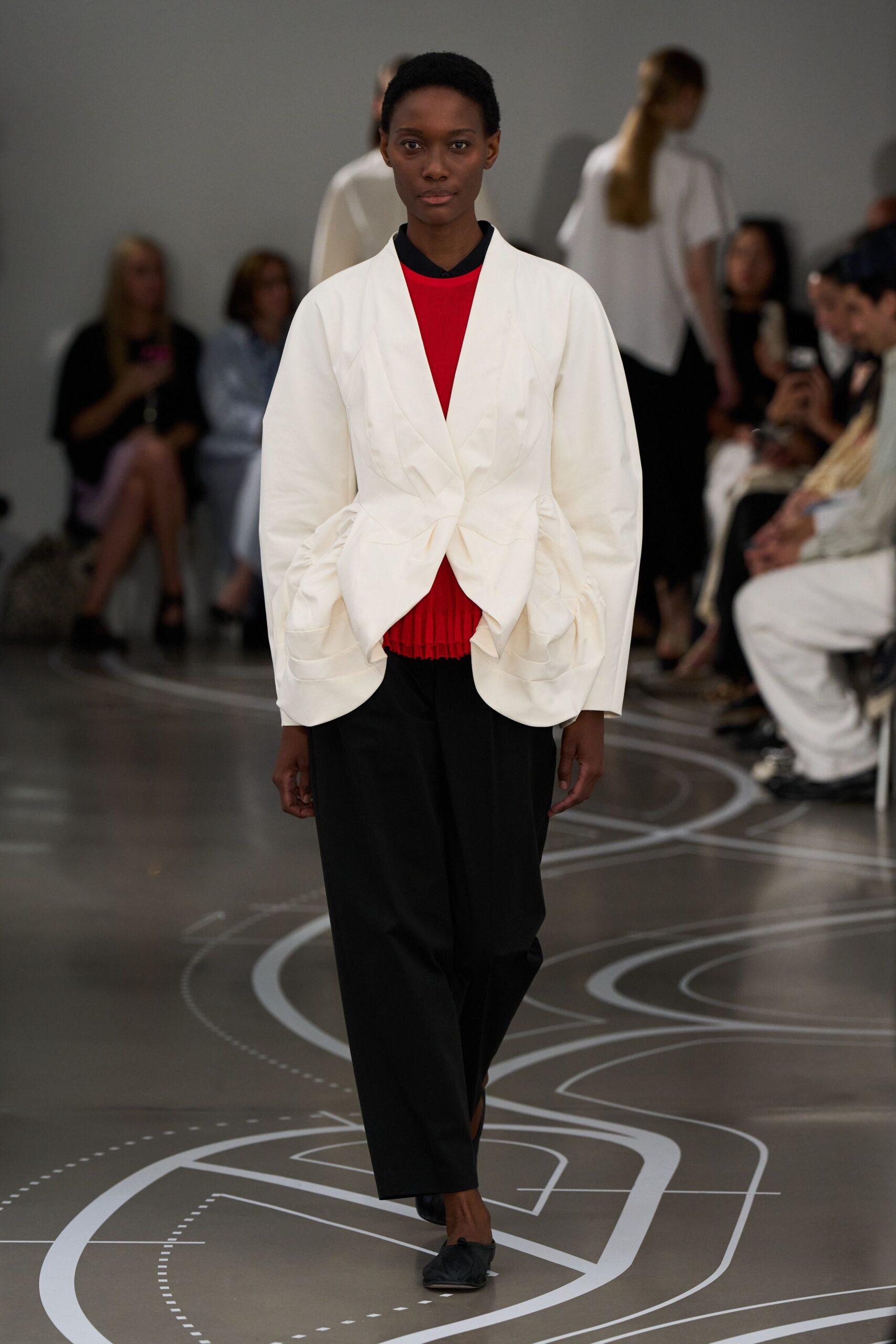Ivory jacket with a twisted front knot and ruched cocoon sleeves. Bright red ribbed knit top, black collar peeking cleanly above. Black wide trousers in soft drape, cropped just above the shoe. Black gathered flats. Crossing an arts campus courtyard after a talk let out early.