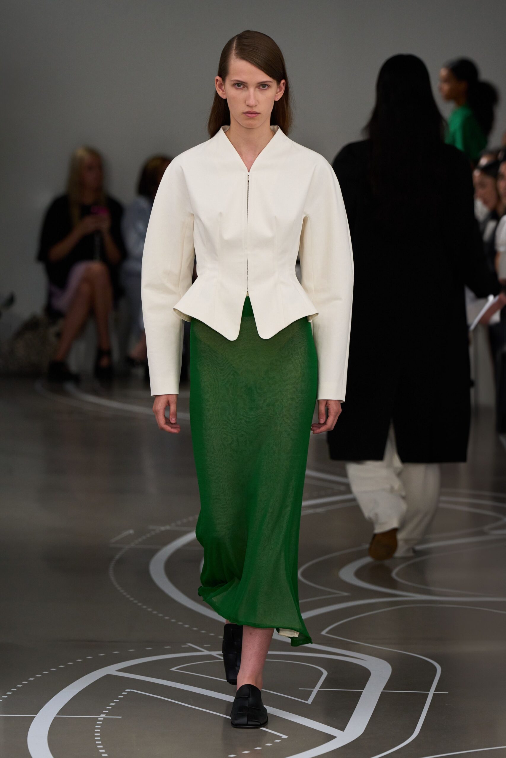 Ivory tailored jacket with a hidden zip and scalloped split peplum. Grass-green sheer knit column skirt, wavering slightly at the ankle. Black woven leather flats. Walking a long museum corridor after the rain has cleared.