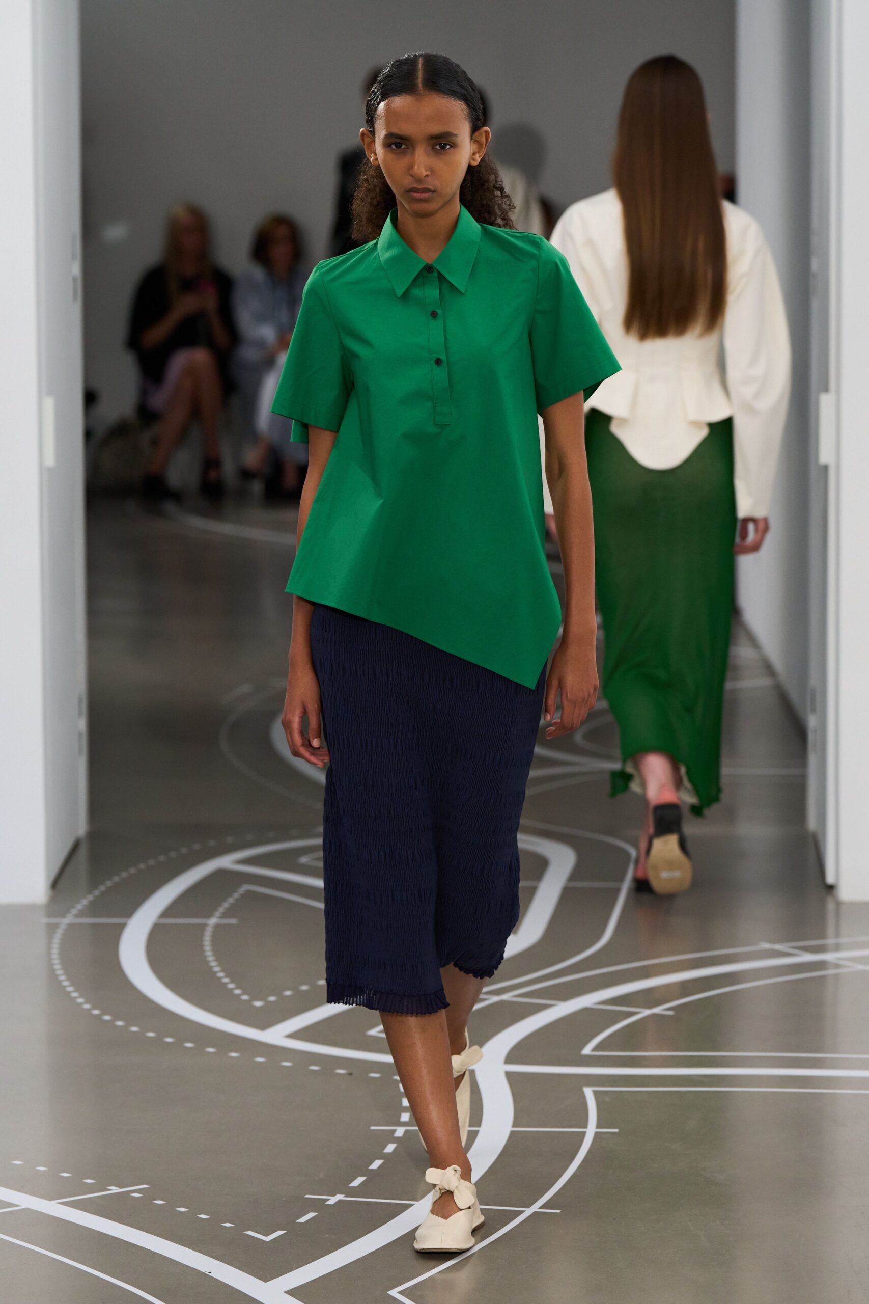 Emerald cotton poplin top with a sharp collar and skewed hem. Navy crinkled knit midi, lightly scalloped at the bottom edge. Cream flats tied with oversized bow straps at the ankle. Walking museum corridors before opening, map lines still bright on the floor.