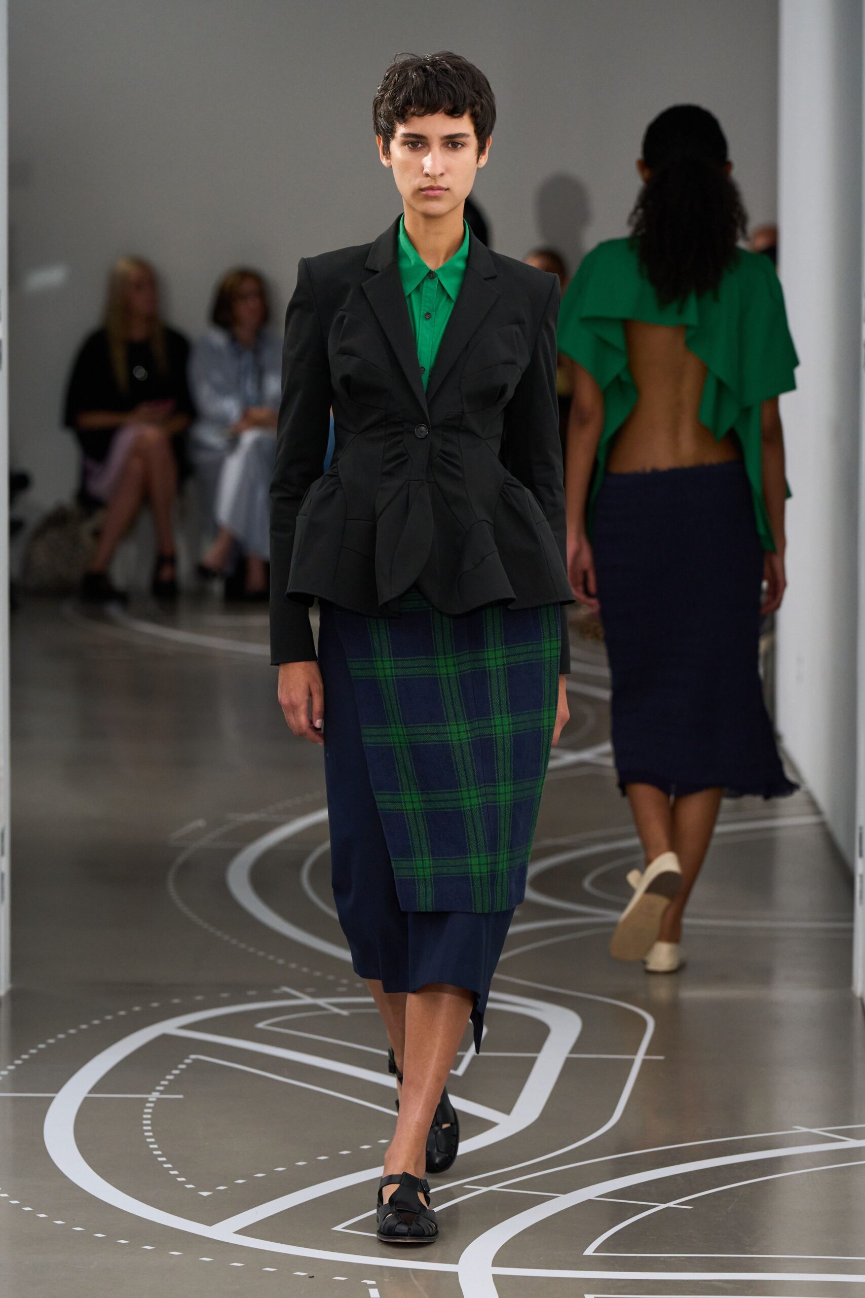 Black tailored blazer, ruched through the waist with padded curved hips. Emerald point-collar shirt, top button fastened under the lapels. Navy wrap skirt with green windowpane panel crossing the front. Black woven leather flats with cutout straps. Walking the long corridor between offices after the building's gone quiet.