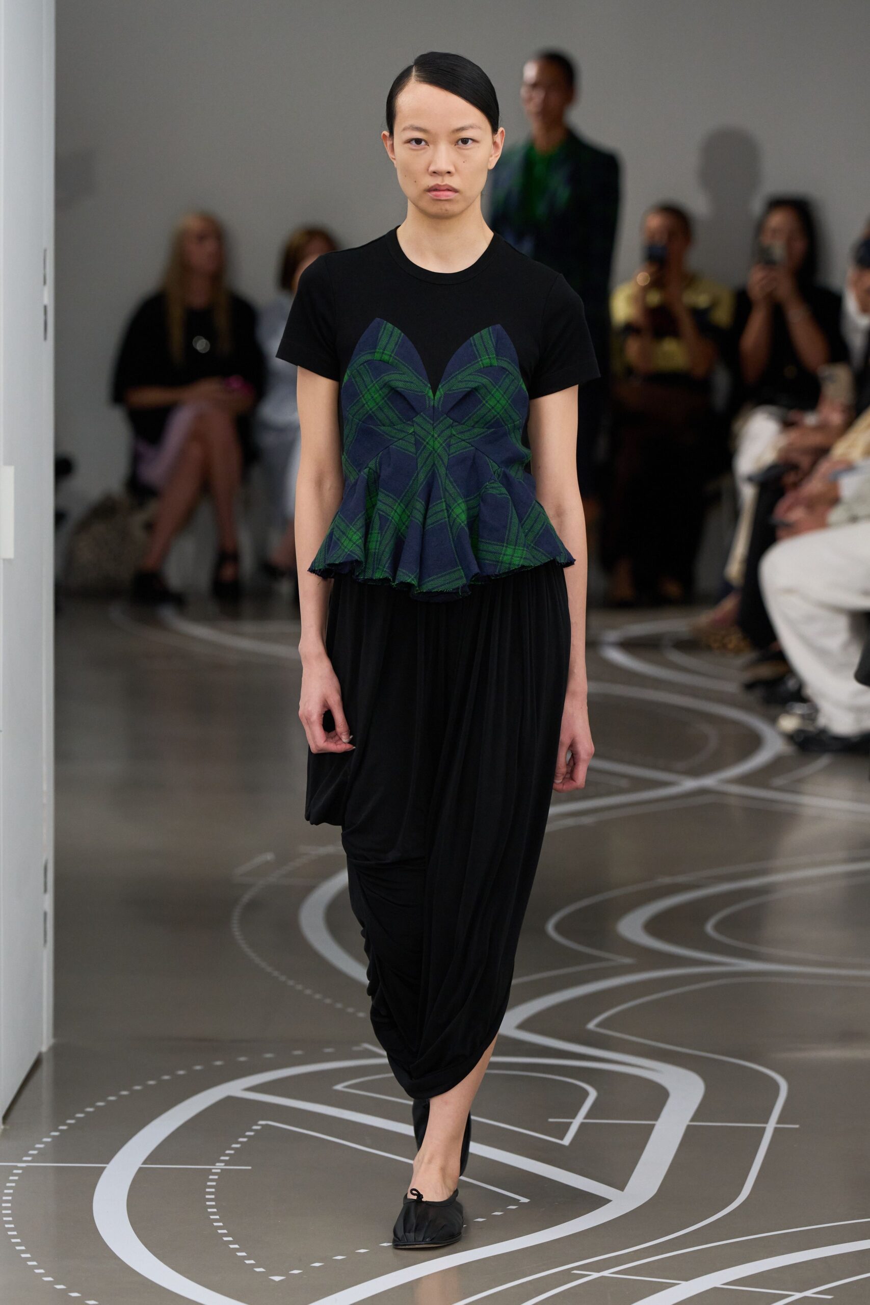 Black jersey tee spliced with a navy-and-green tartan peplum, raw at the hem. Black draped trousers falling in deep jersey folds from the hip. Black satin flats with a tiny bow at the vamp. Crossing a museum atrium after the school groups have gone home.