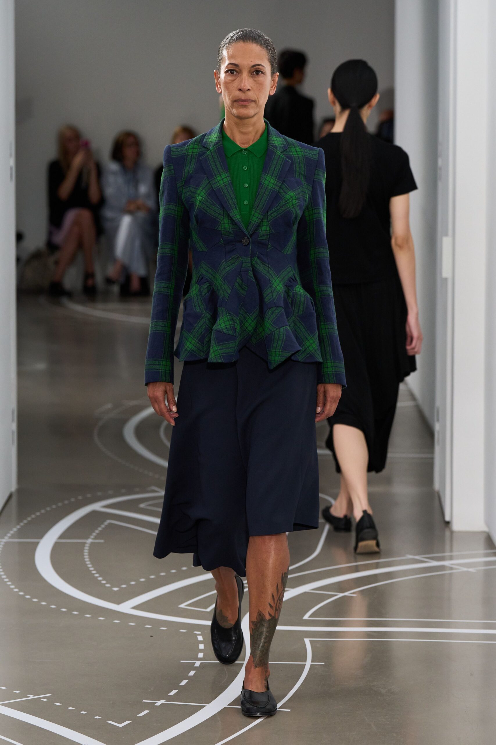 Emerald fine-rib polo, buttoned neatly to the throat. Navy and green tartan jacket, pinched waist flaring into pointed peplum folds. Dark navy skirt falling in an uneven handkerchief hem. Black leather loafers. For crossing a municipal atrium before the public meeting starts.