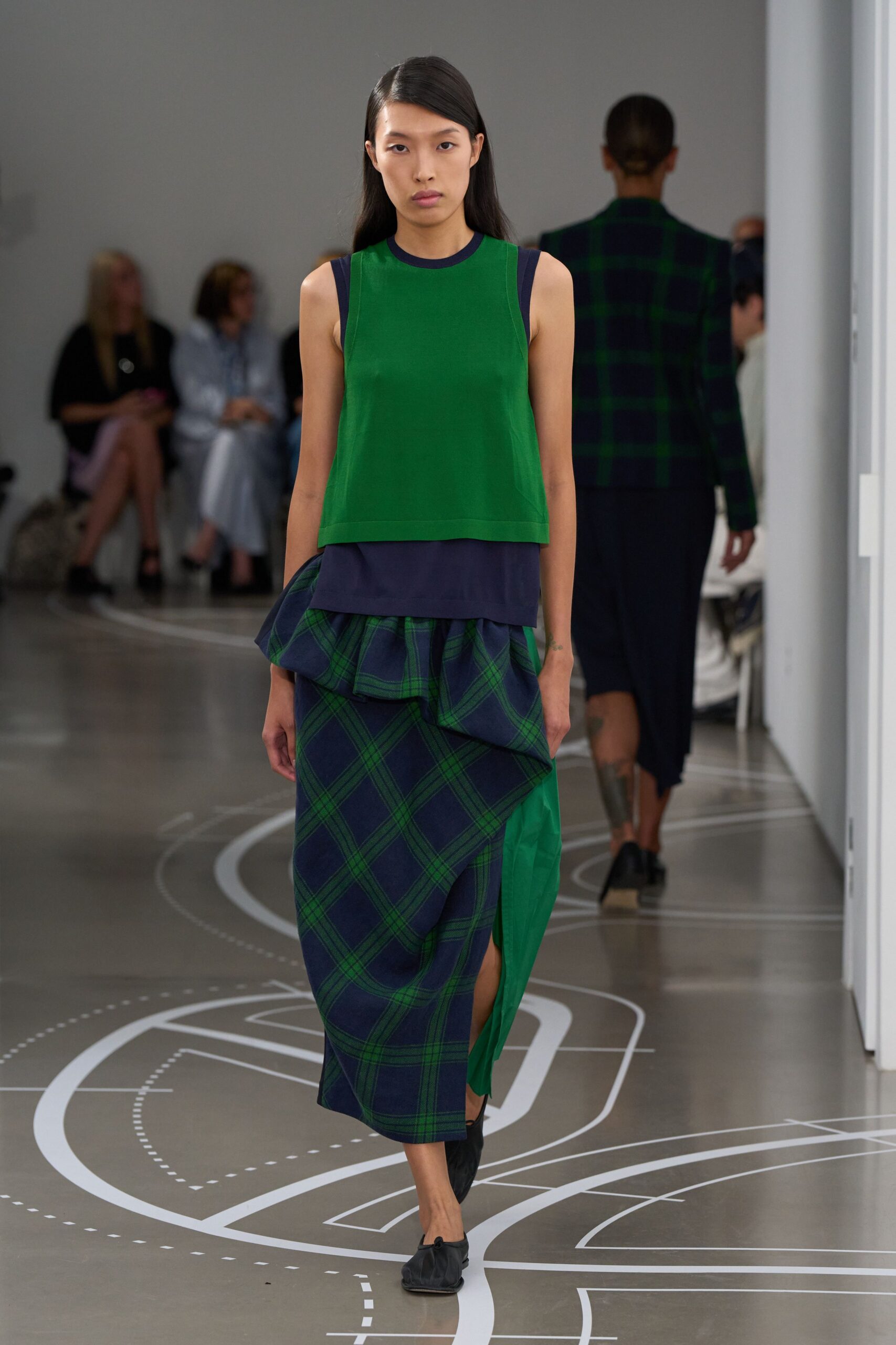 Kelly green knit shell with a navy bound neckline. Longer navy hem peeking below in smooth jersey. Navy and green windowpane wrap skirt, one side in plain green. Black gathered flats. Walking past a campus map after rain, still holding the folded printout.