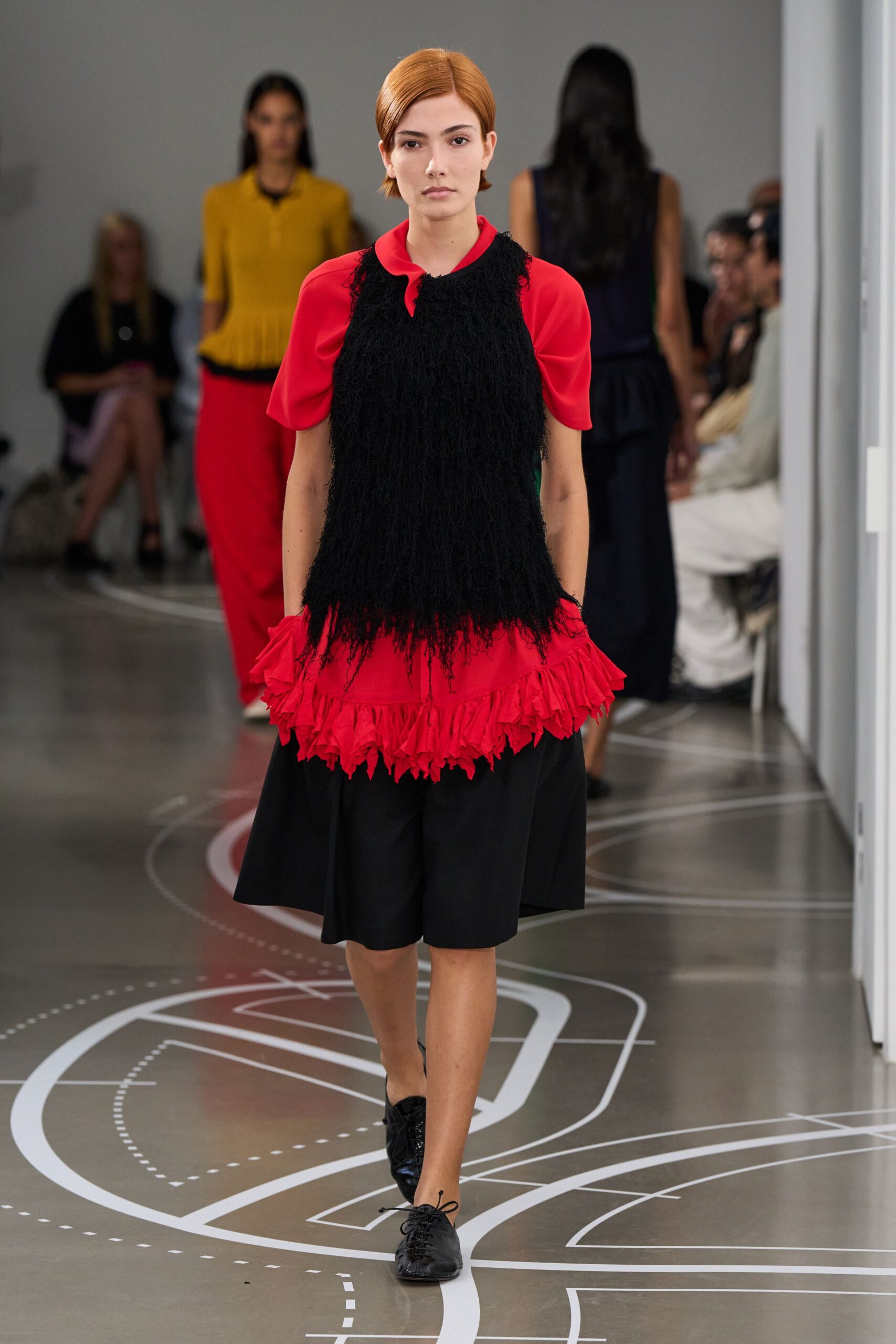 Poppy red dress with puff sleeves and a jagged ruffle hem. Black shaggy fringe layer hanging past the waist in loose strands. Black knee-length culottes cut full and plain beneath the ruffle. Soft black lace-ups, one cord end flicking forward. Crossing a civic atrium after a children's matinee let out.