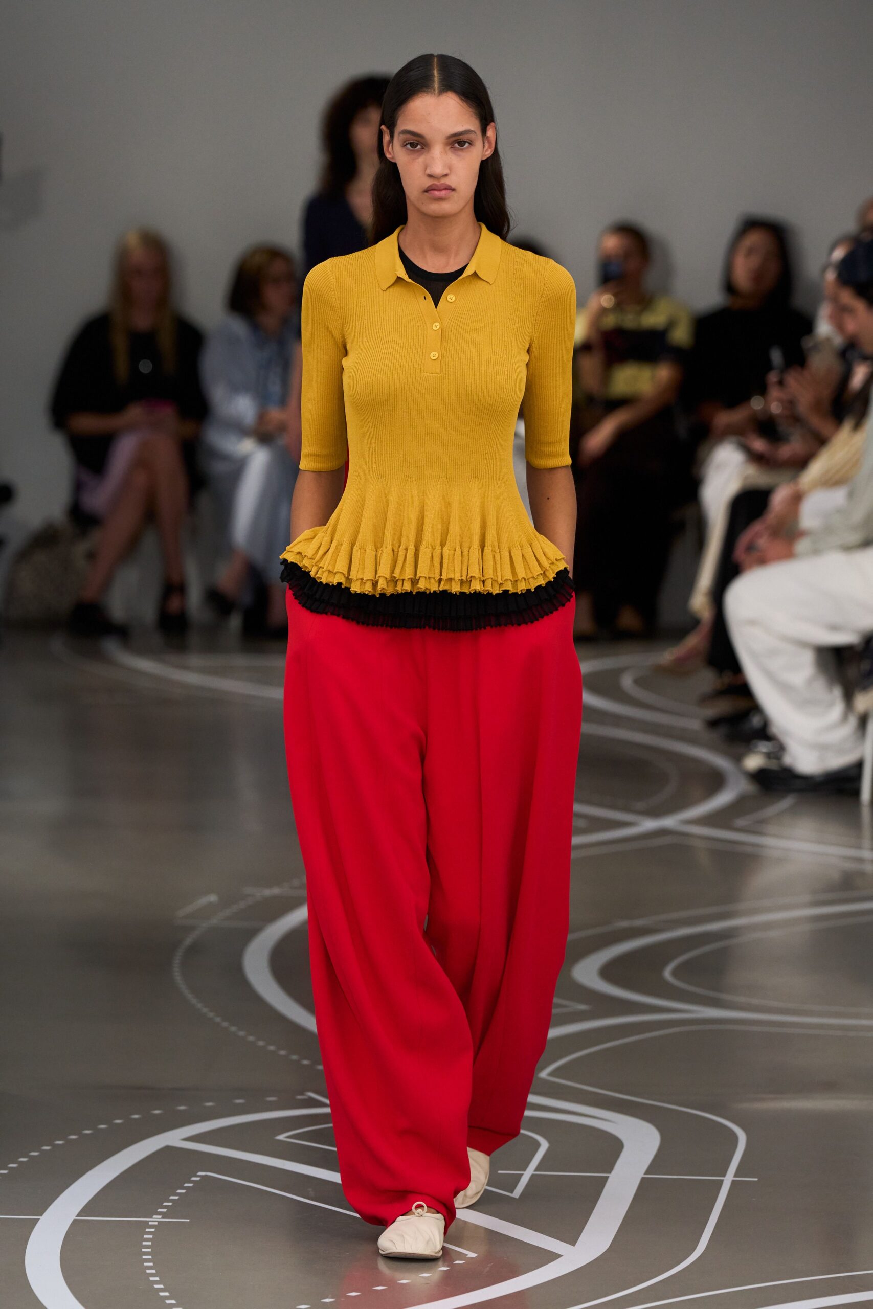 Ochre ribbed knit polo, peplum ending in a tight ruffled edge. Black crewneck insert peeking above the collar and below the hem. Scarlet wide-leg trousers in fluid crepe, breaking softly over the shoe. Cream square-toe flats with a gathered vamp ring. Lunch on a museum terrace after you missed the train south.