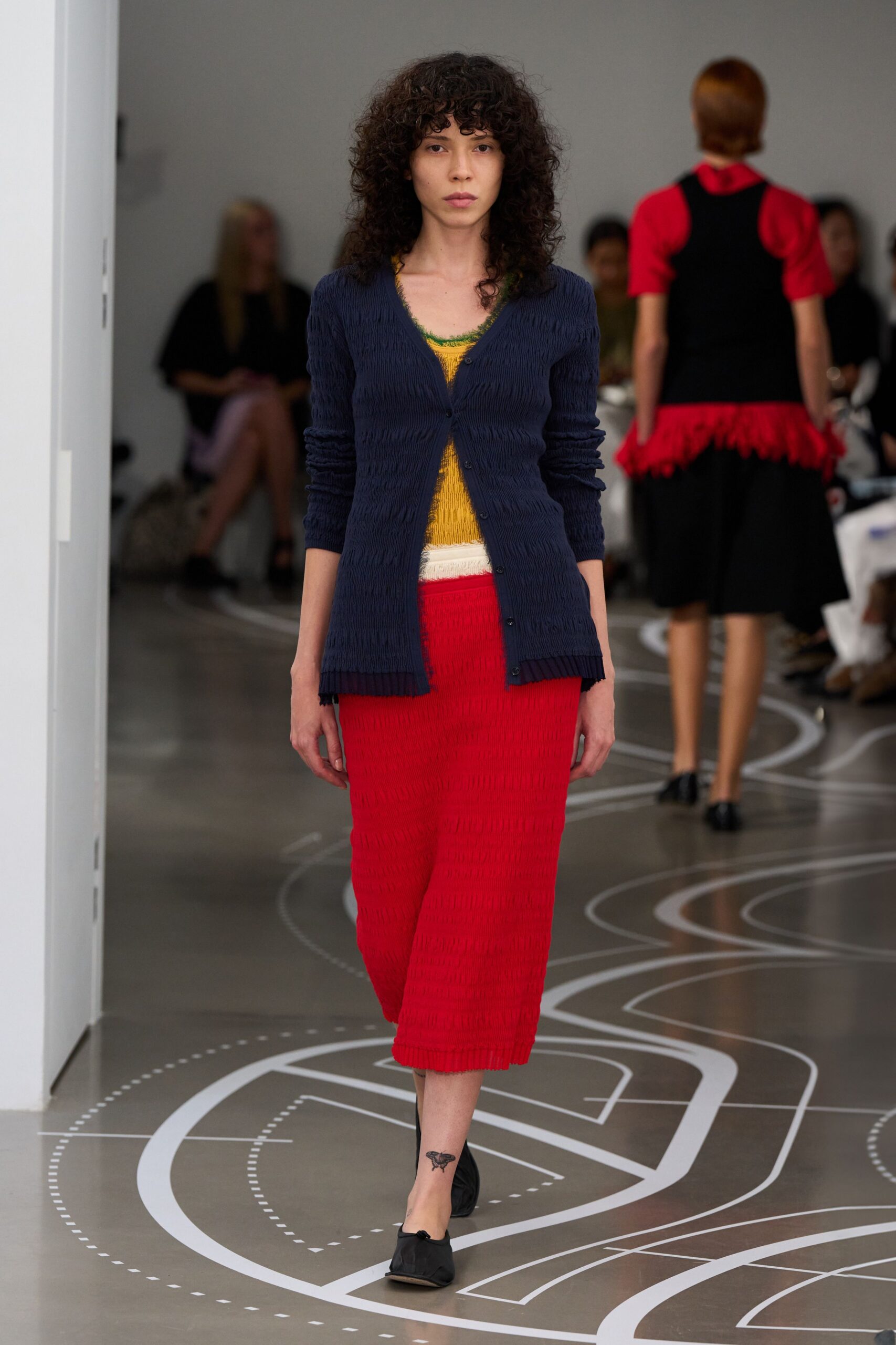 Navy crinkled cardigan, narrow placket skimming open over the torso. Mustard ribbed knit top with a moss-green scalloped neckline. Bright red crinkled midi skirt with a chalk-white band at the waist. Black folded flats with soft squared toes. Walking the long way home after buying peaches at the Saturday market.