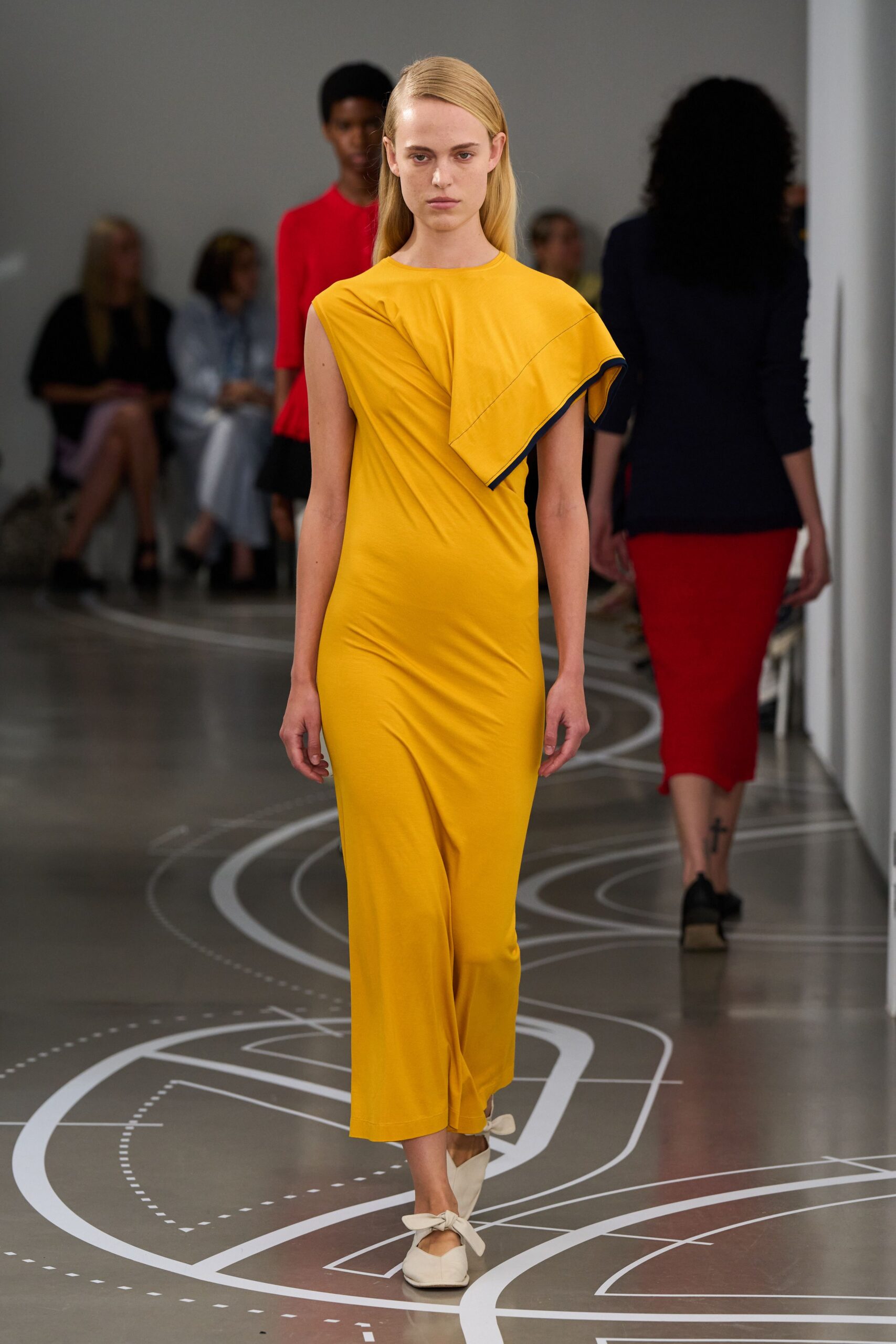 Marigold jersey column dress, one shoulder folded into a navy-edged capelet. Cream pointed flats tied with oversized bows across the instep. Crossing a ferry terminal with flowers wrapped in yesterday's paper.