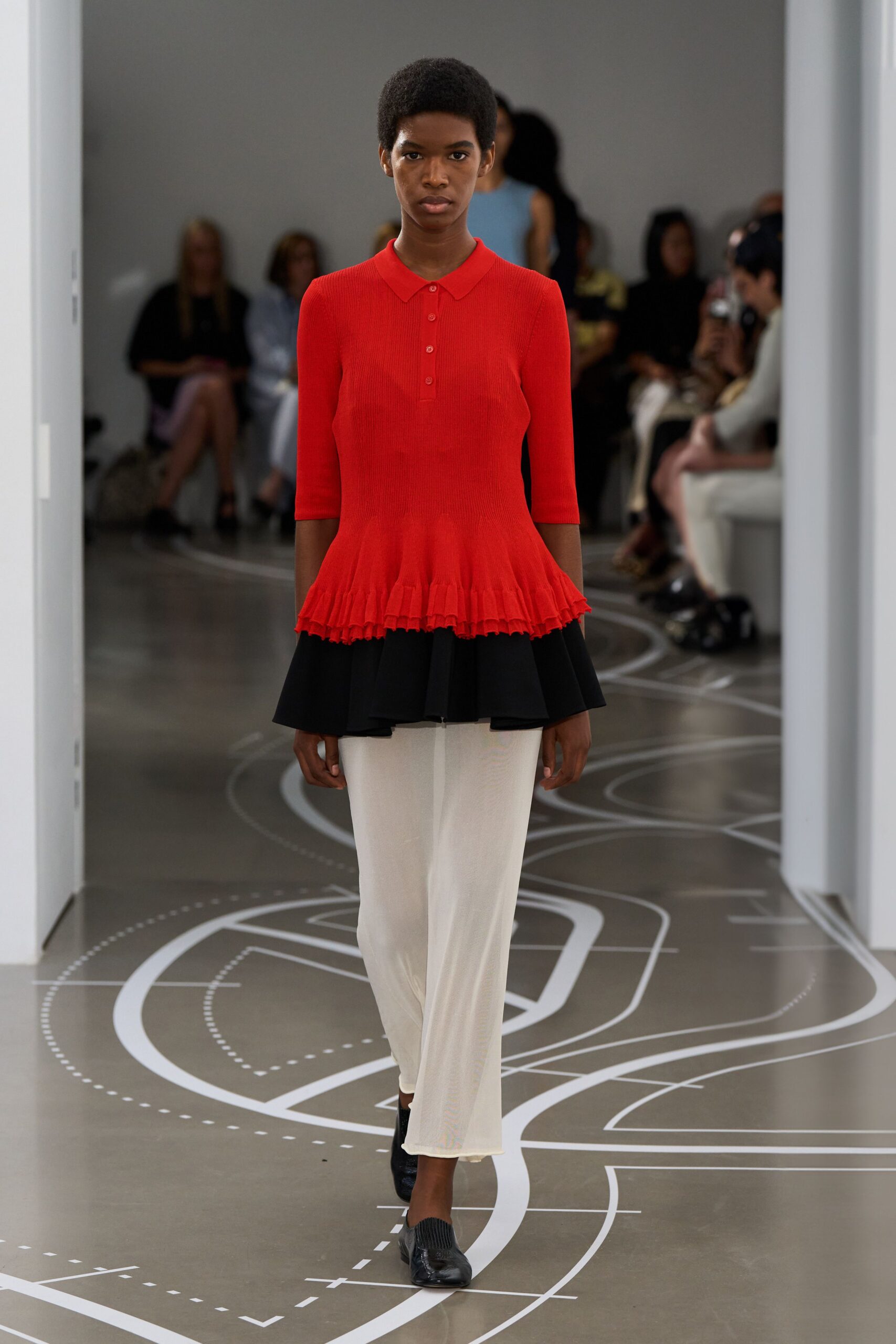 Tomato red rib knit polo, button placket ending above a fluted peplum. Black circular flounce layered under the peplum, swinging past the hips. Ivory column skirt in a fine translucent knit. Black square-toe flats with ribbed vamp panels. Crossing a civic atrium before the building opens, footsteps loud on concrete.