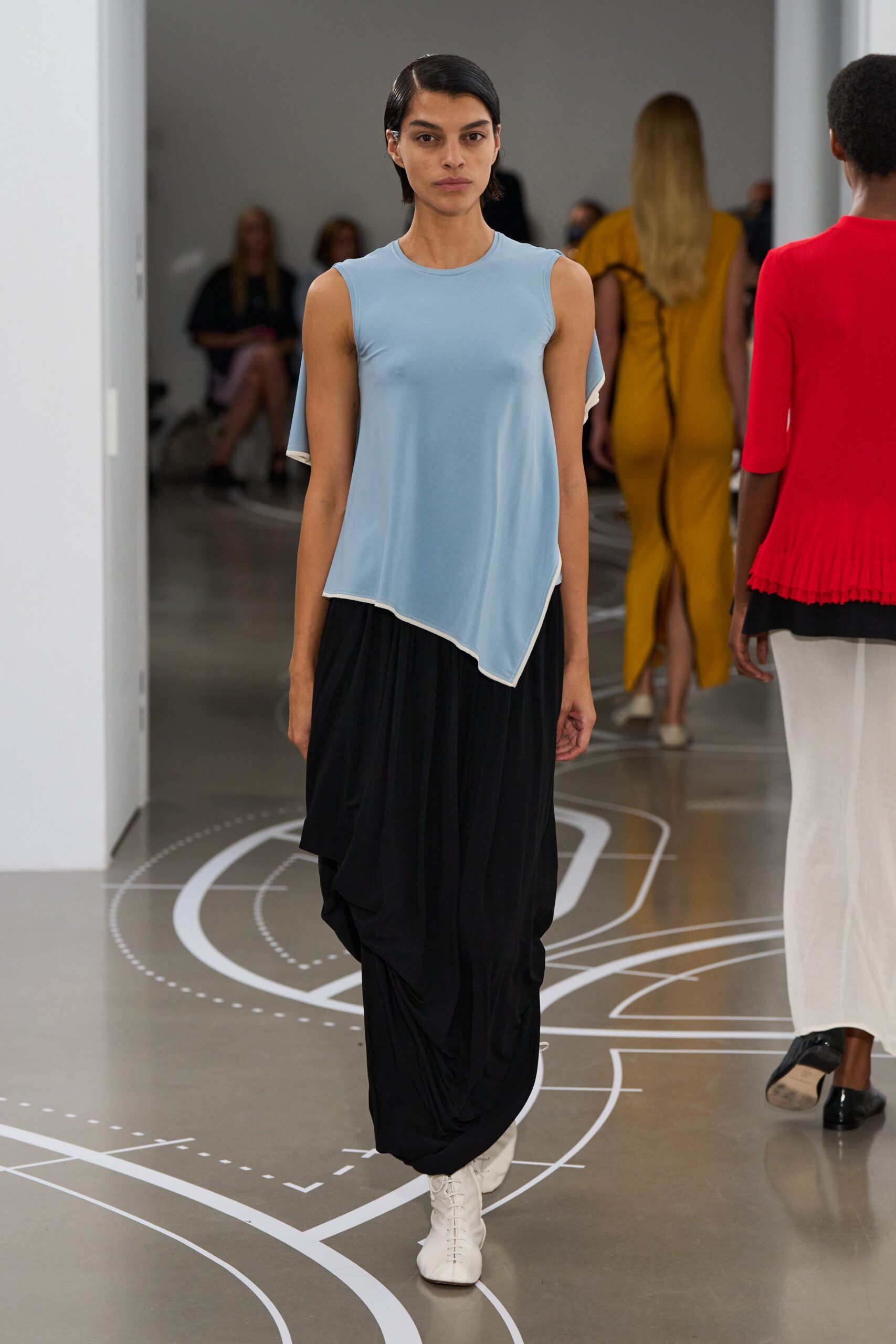 Powder-blue jersey top with a sharp handkerchief hem edged in white. White knit sleeve peeking from the armhole on one side. Black draped skirt gathered into deep hanging folds. White lace-up ankle boots with a squared-off toe. Crossing a museum atrium while the floor plan curls underfoot.