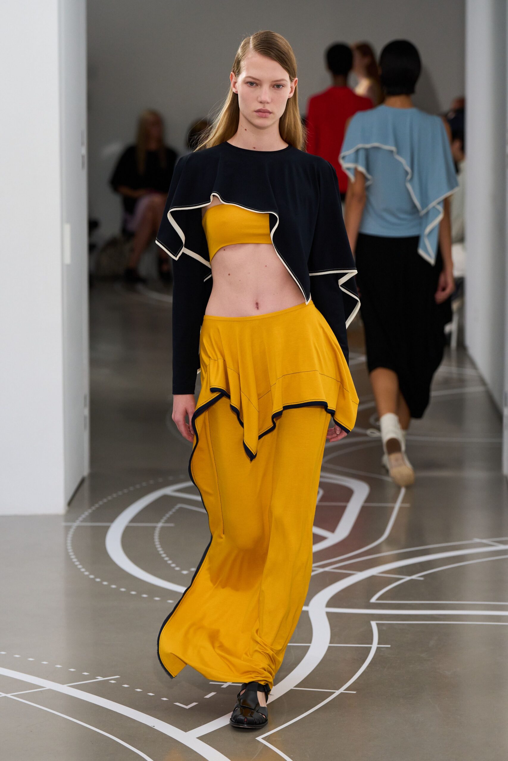 Black cropped cape top with white binding tracing the ruffled edge. Marigold bandeau in smooth stretch jersey, cut straight across. Marigold jersey skirt-trousers with an uneven peplum and ink-dark piping. Black fisherman sandals with broad cutout straps. Crossing a sunlit atrium with museum plans painted underfoot.