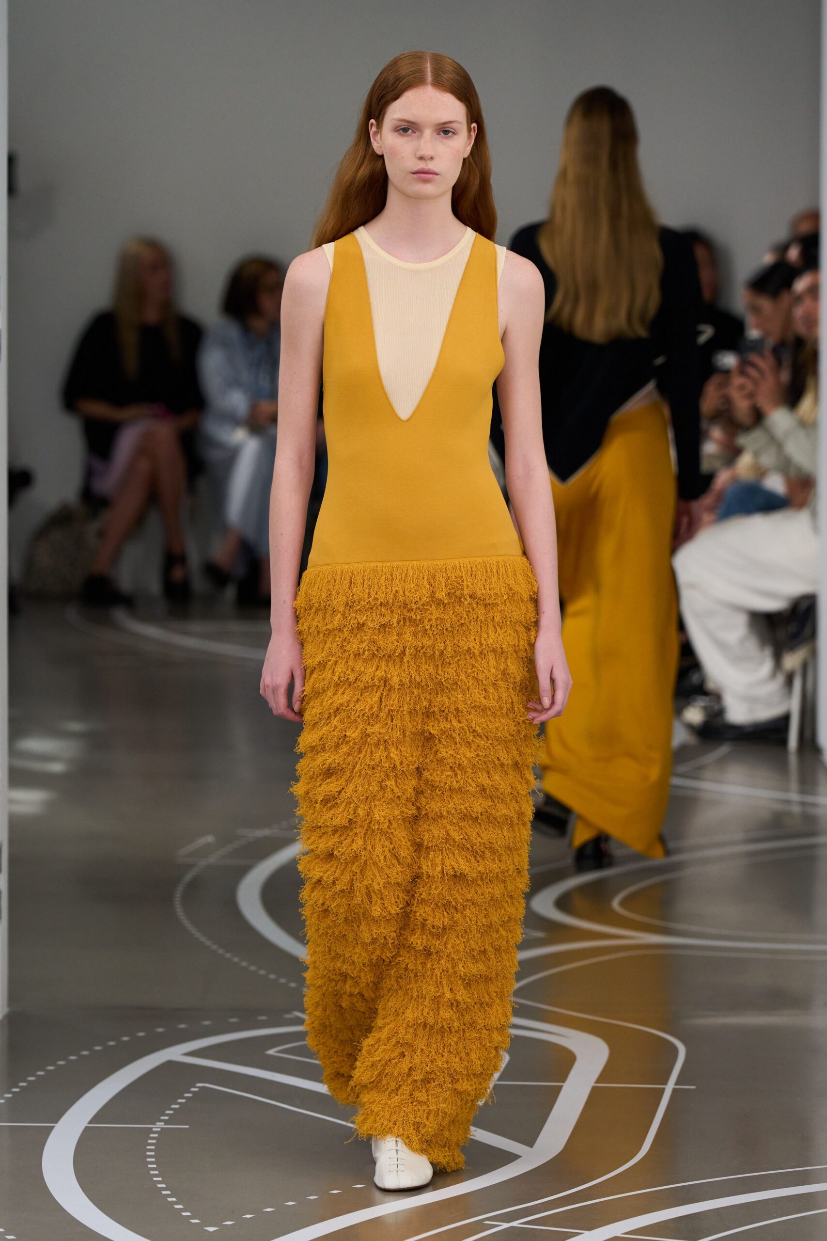 Marigold column dress with a sheer cream inset plunging to the sternum. Dense shag fringe builds from the dropped waist to the floor. White square-toe shoes with a narrow split up the vamp. Crossing an echoing museum atrium after the rain has stopped.
