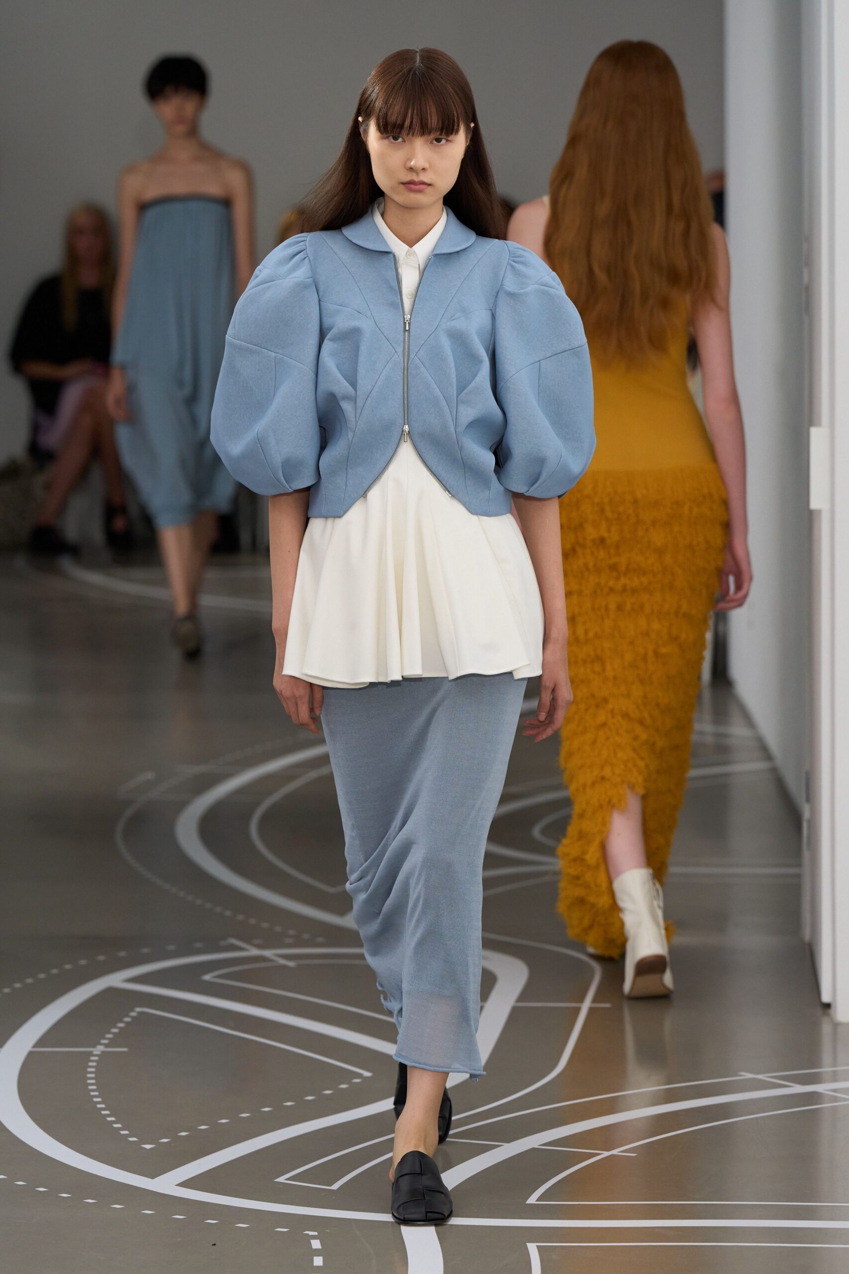 Powder-blue cropped jacket with quilted seams and swollen puff sleeves. White pointed-collar shirt falling into a soft peplum underlayer. Matching blue jersey column skirt, hem rolling slightly at the edge. Black square-toe flats. Walking a quiet office corridor before anyone switches on the lights.