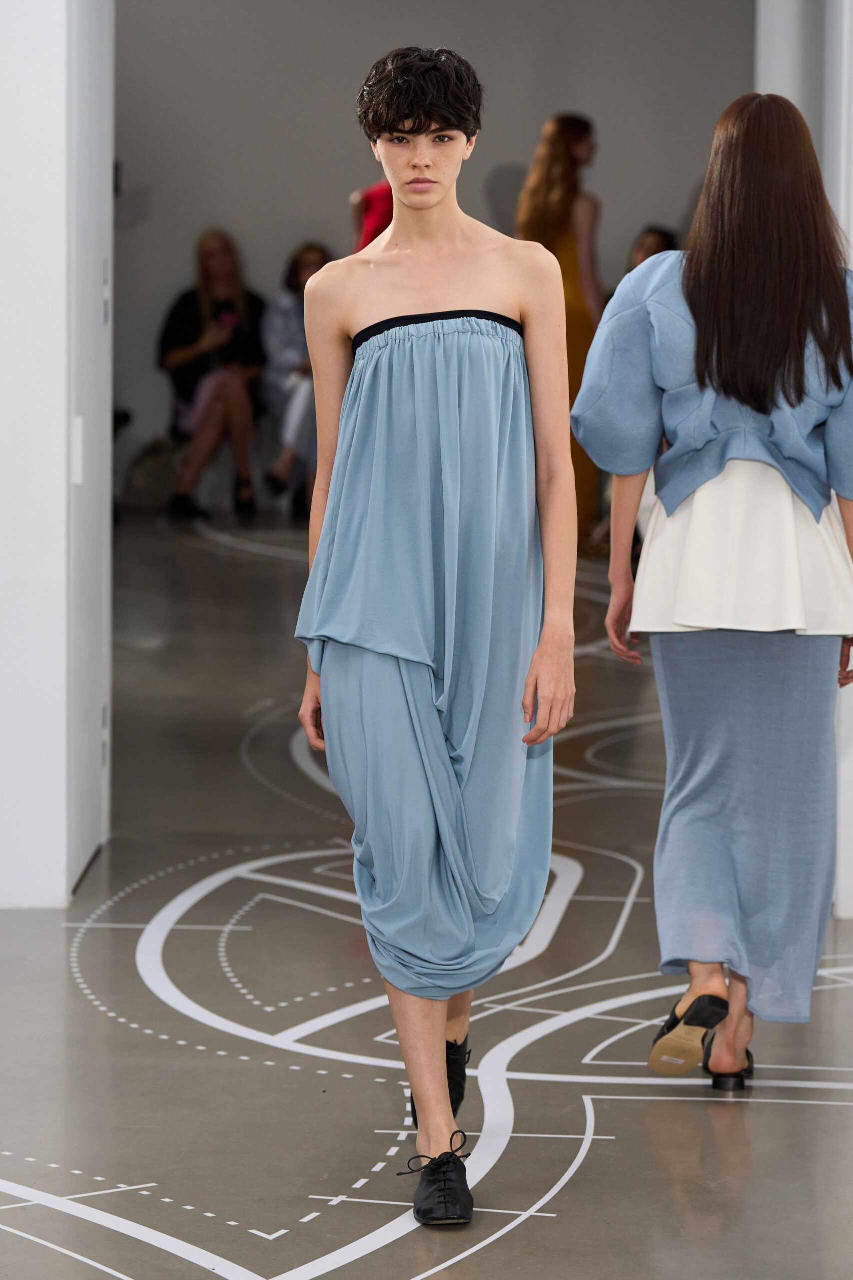 Powder-blue jersey tube dress, black binding at the gathered top edge. Black lace-up flats with a narrow squared toe. Crossing a museum atrium before the air-conditioning fully kicks in.