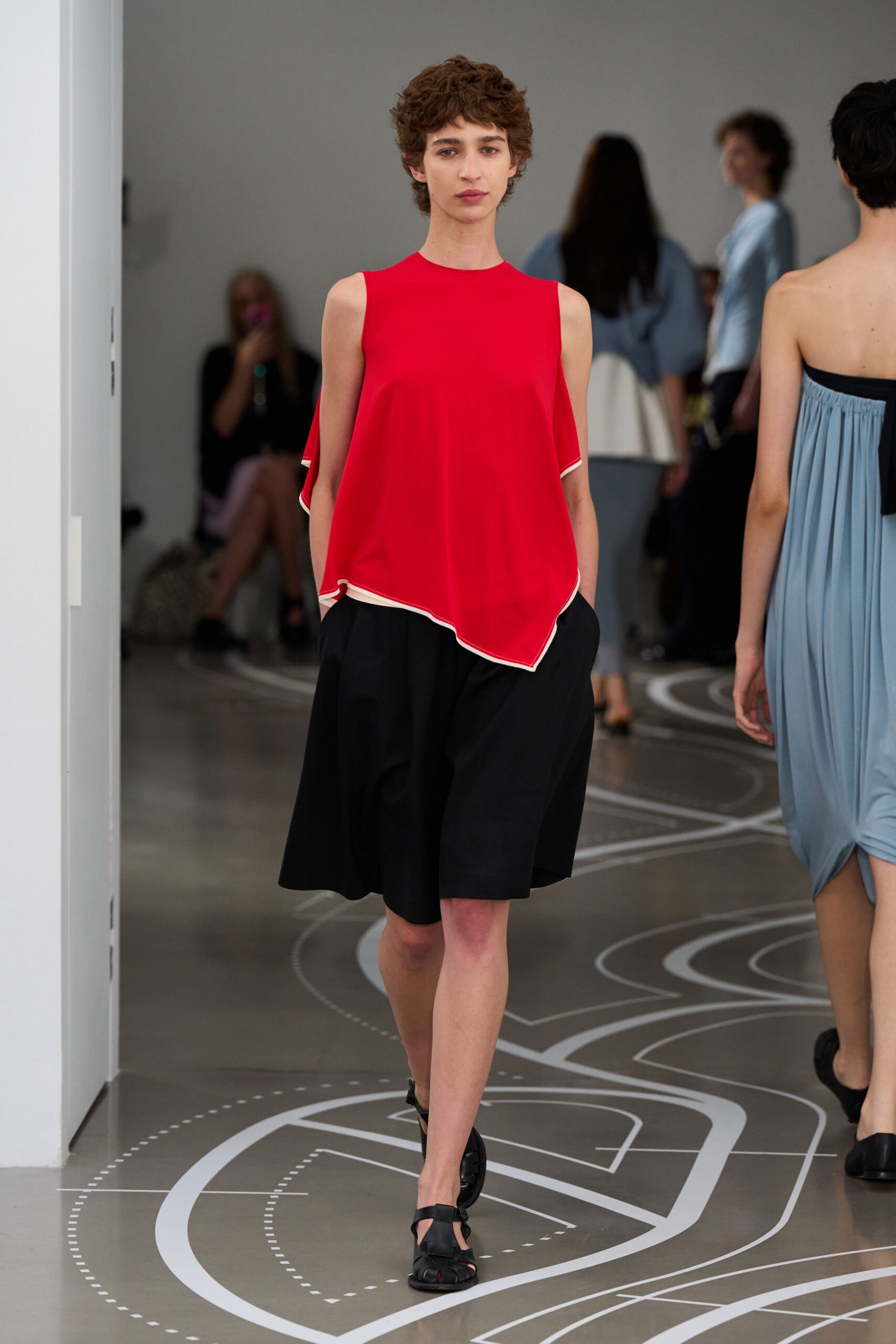Scarlet draped shell top with white rolled edging and a pointed handkerchief hem. Black knee-length culottes cut wide, hands sunk into side pockets. Black cutout sandals with a T-strap front. Crossing a museum atrium during the quiet hour before school groups arrive.