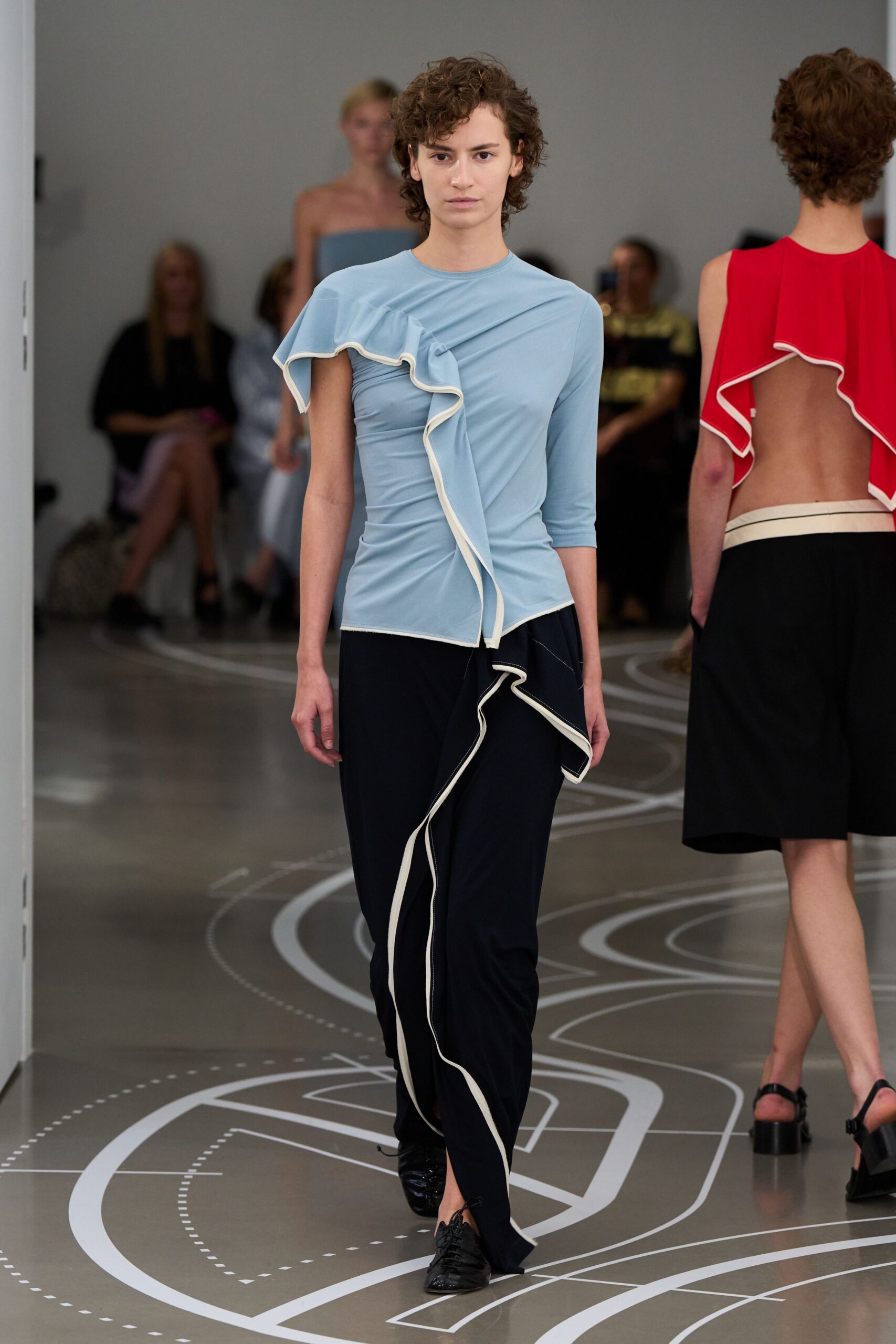 Powder-blue stretch top, one sleeve cut away under a cream-edged ruffle. Black fluid trousers with a cream-bound flounce twisting from hip to hem. Black lace-up flats, softly creased at the toe. Crossing the museum atrium before the school groups arrive.