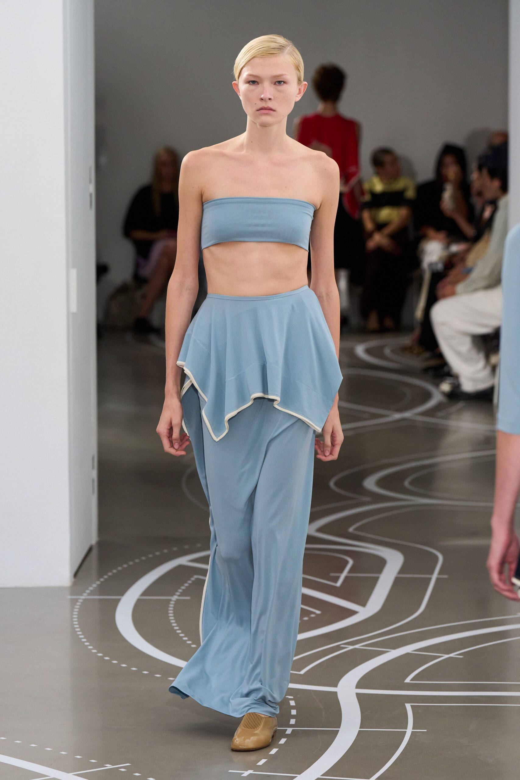Powder-blue stretch bandeau, cut straight and held close to the ribcage. Liquid jersey trousers with a handkerchief peplum edged in white binding. Tan leather flats with ribbed inserts across the instep. Walking the tiled concourse before the aquarium opens.