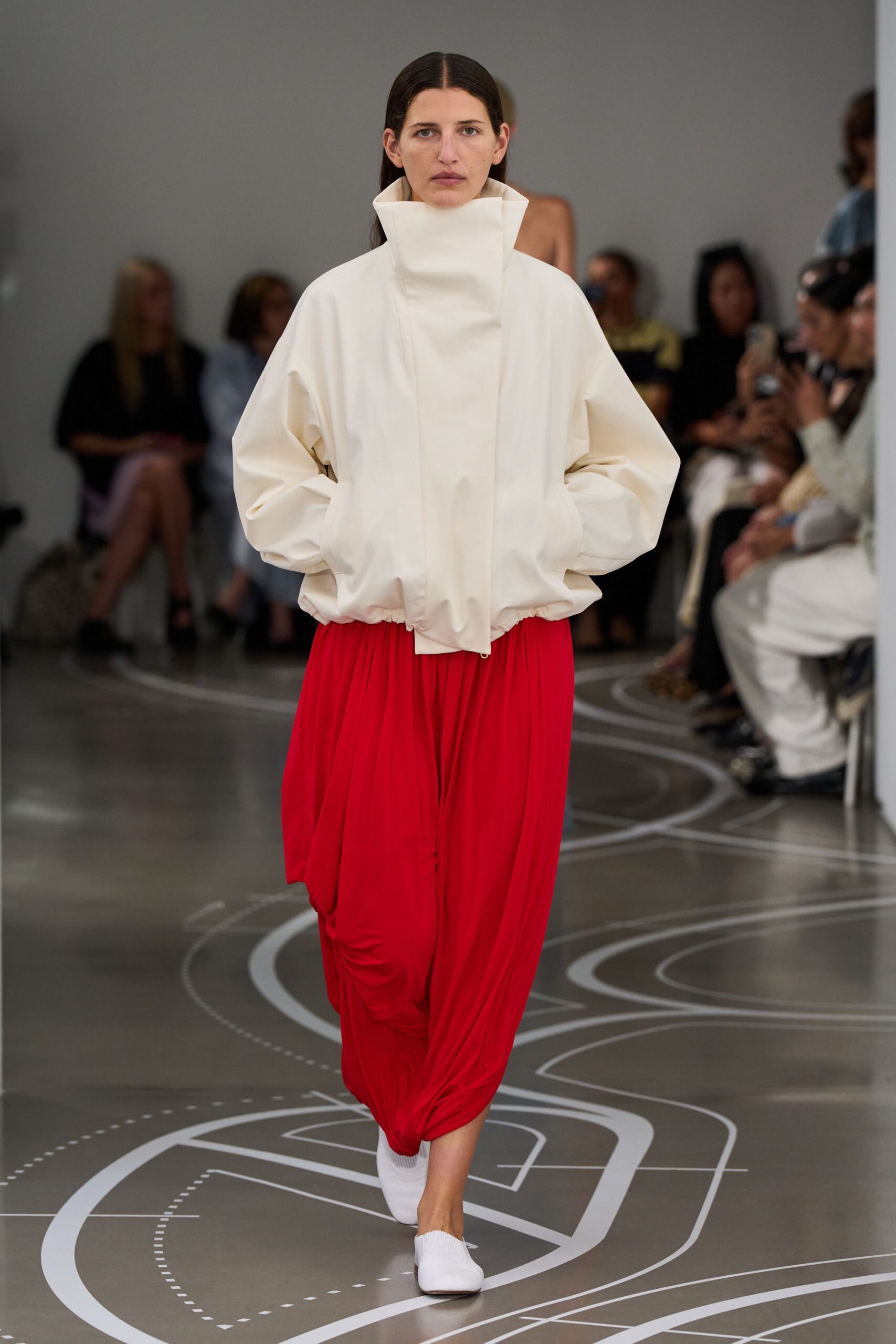 Cream poplin jacket with a tall funnel collar and elasticated hem. Red jersey draped into a low slung cocoon, gathered at one knee. White knit sock shoes with blunt squared toes. Crossing a museum atrium before the rain starts.