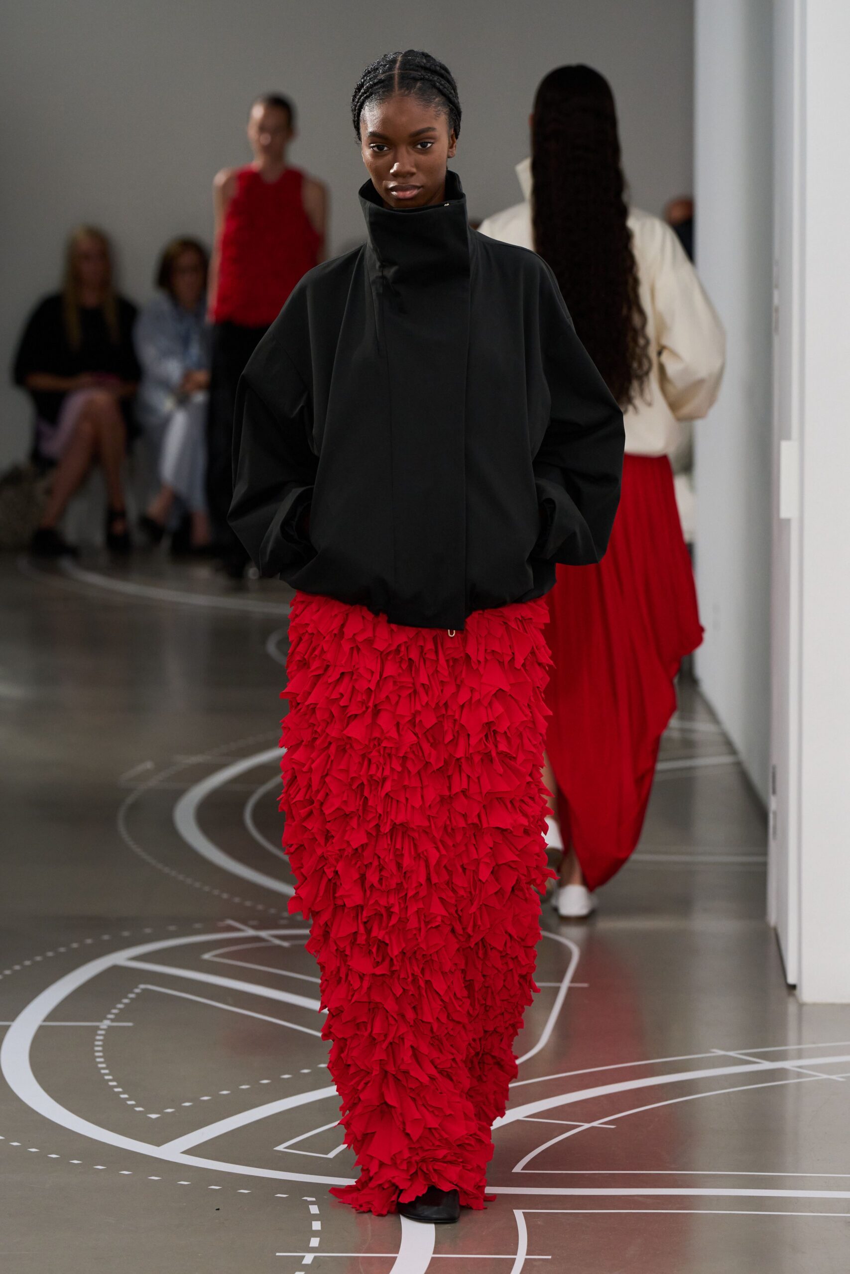 Black cocoon jacket with a high funnel neck and bloused hem. Floor-length scarlet skirt buried in dense raw-edged ruffles. Black square-toe boots peeking out below the frayed red sweep. Crossing a museum atrium while a school group scatters ahead.
