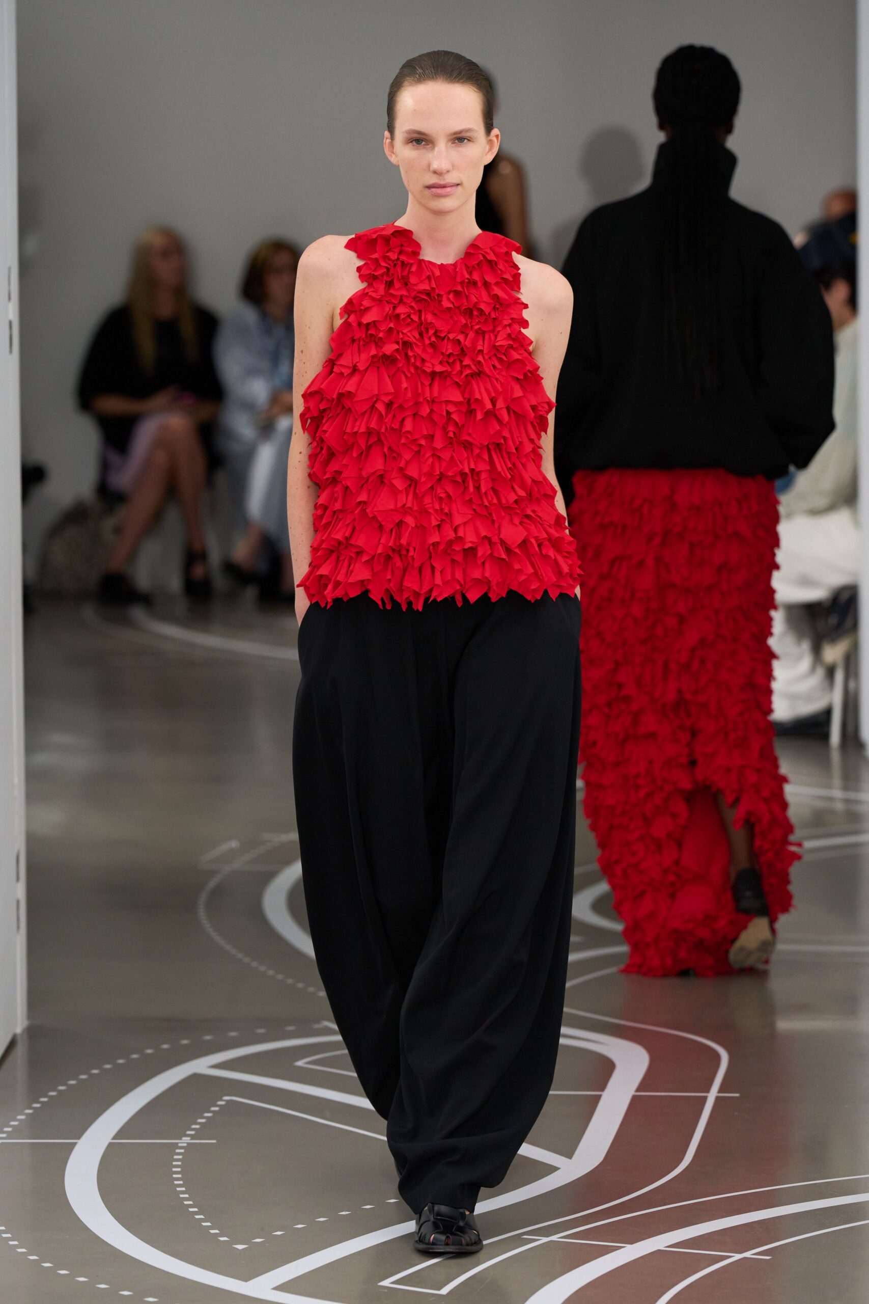 Scarlet sleeveless top built from dense fluttering ruffles, hem left jagged. Black fluid trousers cut full through the leg, hands sunk in pockets. Black caged sandals. Crossing a museum atrium after the florist buckets have just been tipped out.