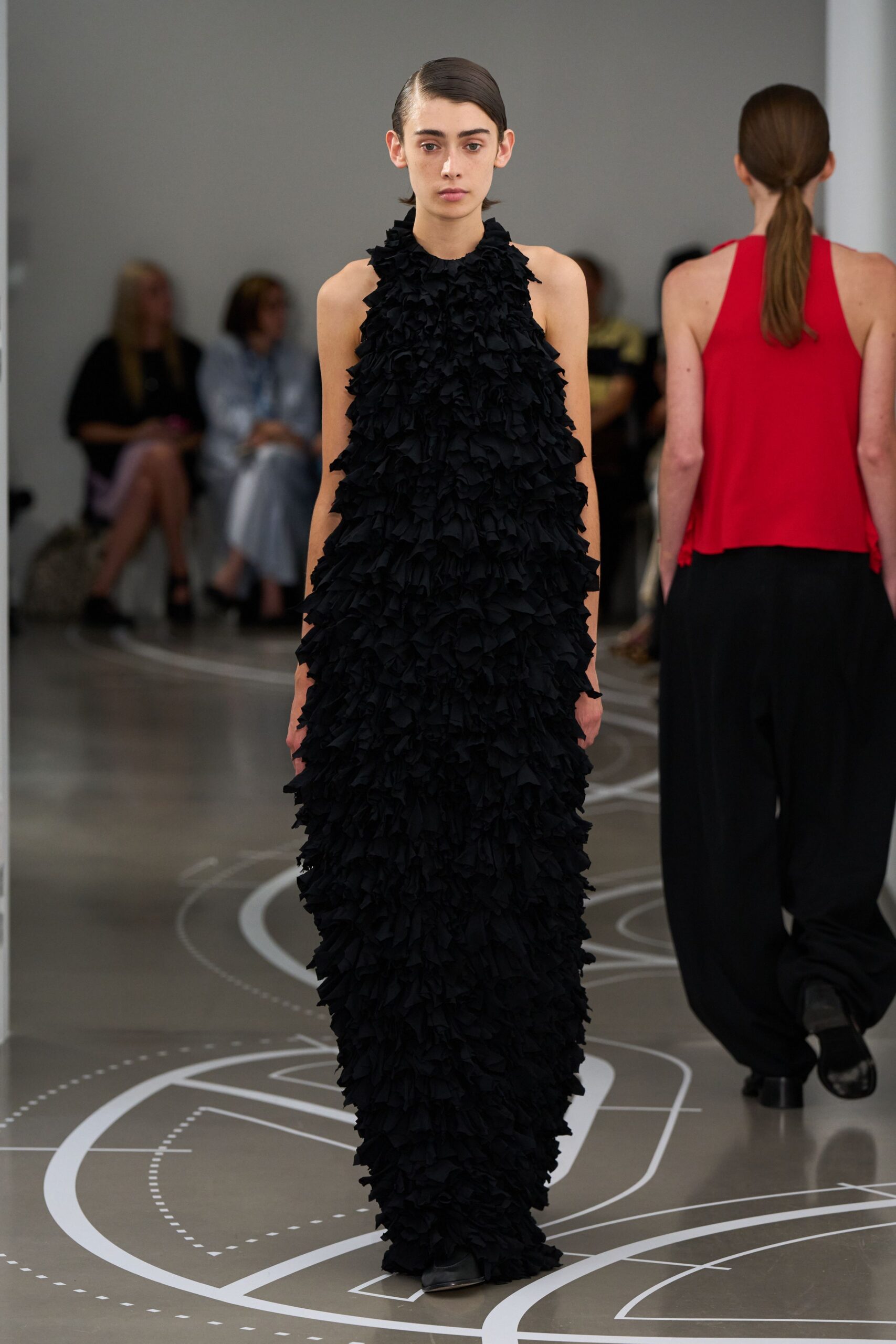Black halter-column gown buried under dense shredded ruffles from throat to hem. Black square-toe shoes peeking from the pooled edge. Crossing a museum atrium while the floor plan curls underfoot.