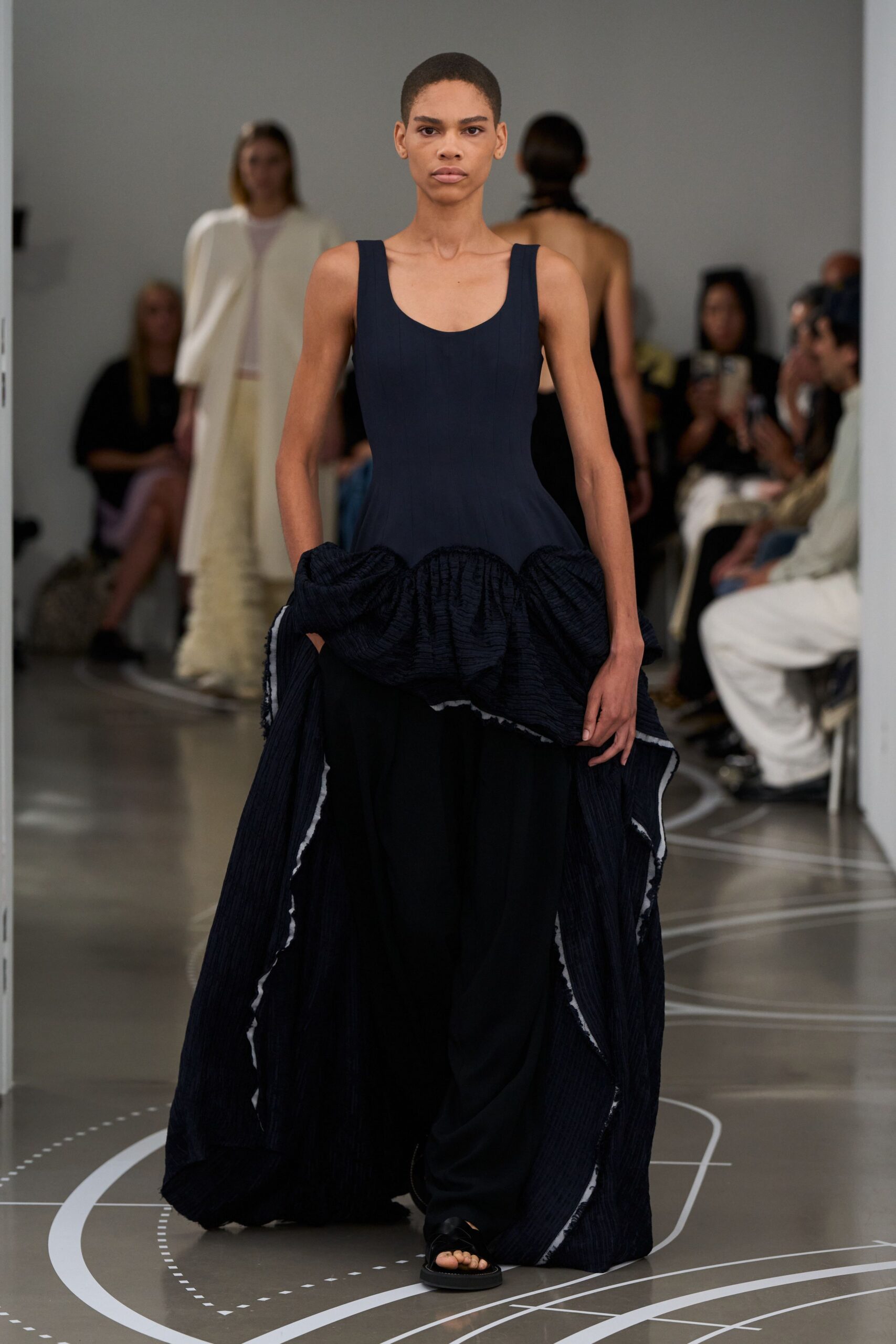 Ink-black sleeveless dress with a scooped neckline and ruched bubble peplum. Wide black trousers veiled by ribbed panels edged in stark white. Black slide sandals with a broad padded strap. Crossing a museum atrium after rain, floor still marked for the installers.