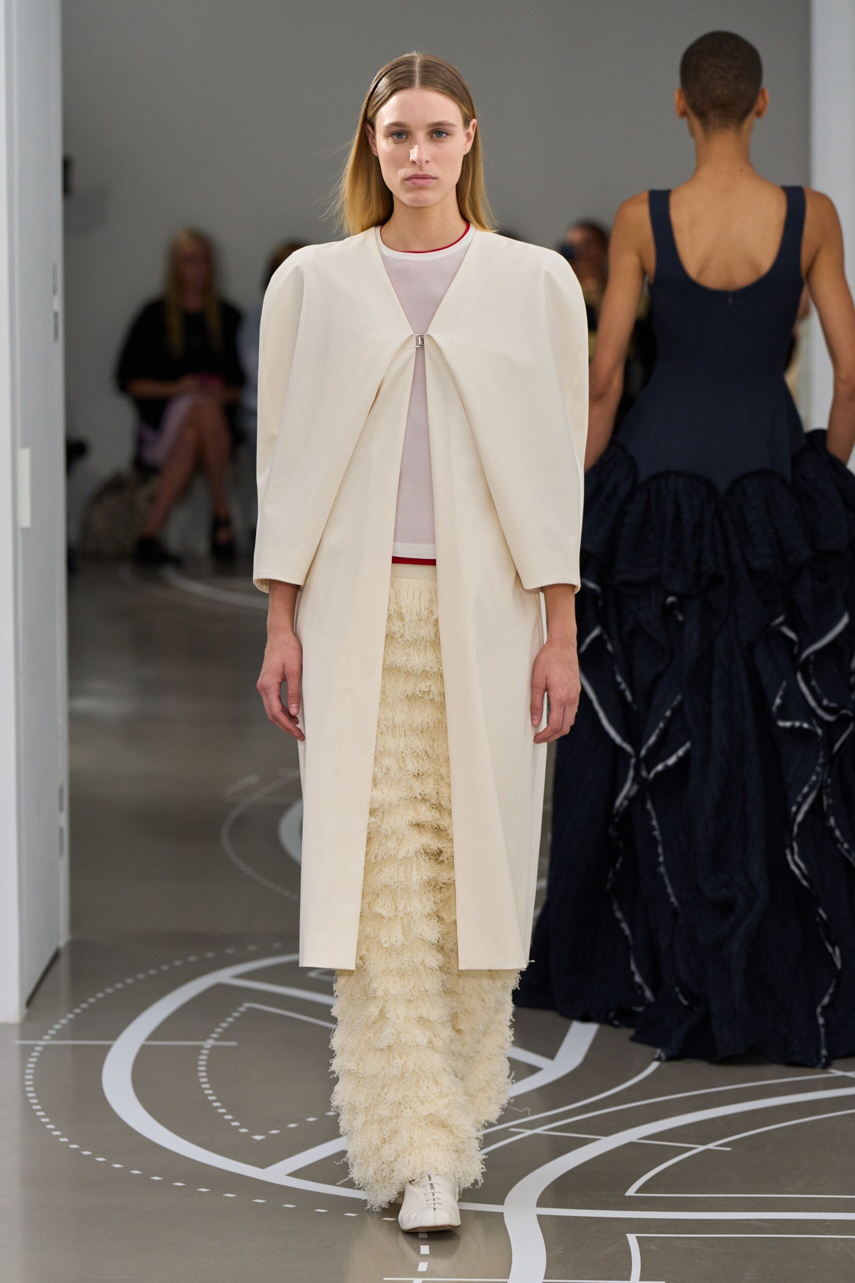 Ivory cocoon coat with a single clasp pinching the front pleat. Sheer white mesh shell, outlined at neck and hem in red. Long cream skirt built from shaggy tiered fringe. White square-toe boots with center-front lacing. Museum atrium at noon, footsteps echoing off the stone floor.