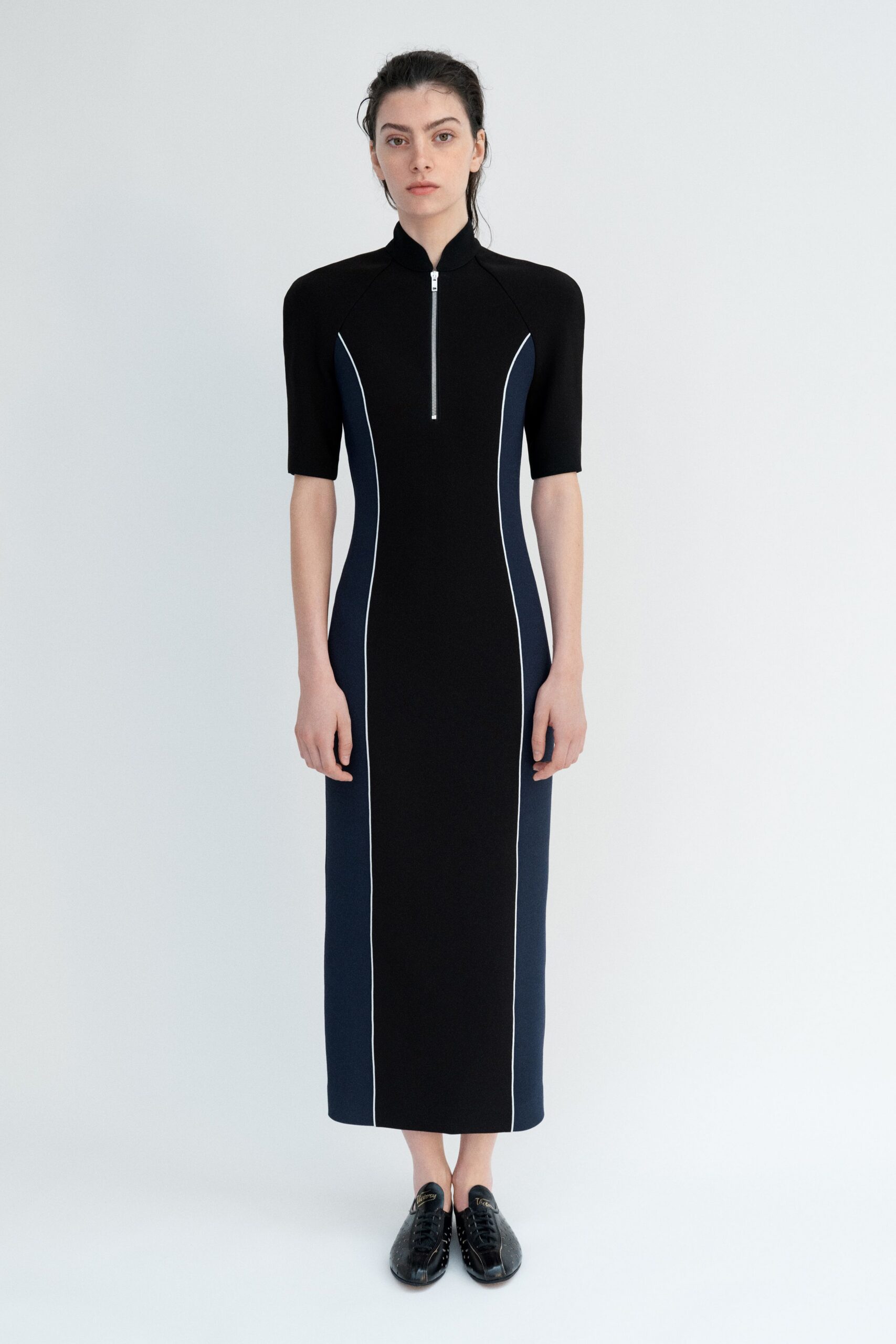 Black knit column dress with navy side panels and white piping. Half-zip ending mid-chest beneath a close stand collar. Black leather lace-ups with a pale seam splitting the toe. Walking the long museum corridor before the building opens.