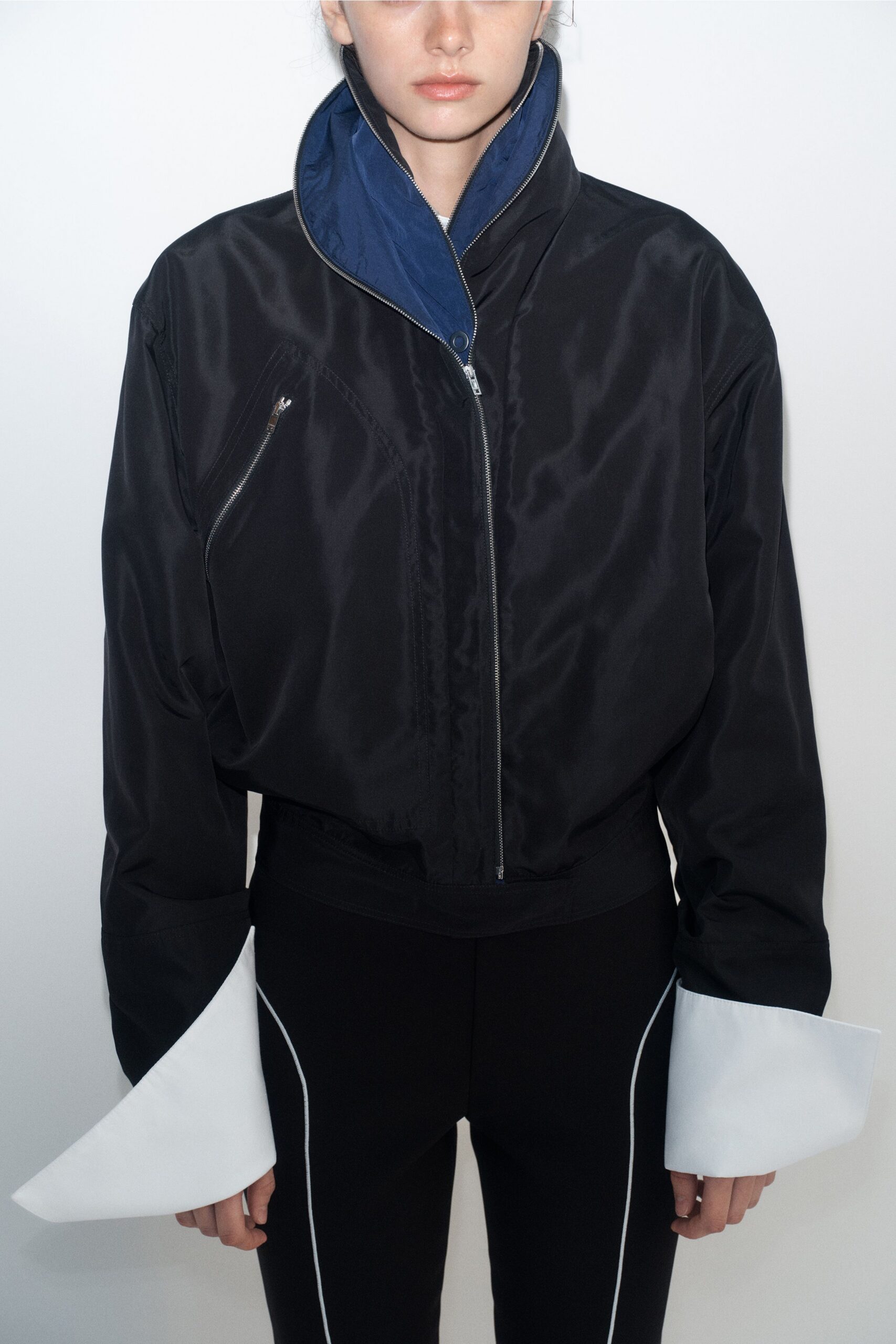 Black satin bomber with cobalt stand collar and a slanted chest zip. Oversized white cuffs, stiff and flared like folded card. Black slim trousers traced with curved white piping at the outer leg. Walking a windy ferry deck with your ticket already damp.