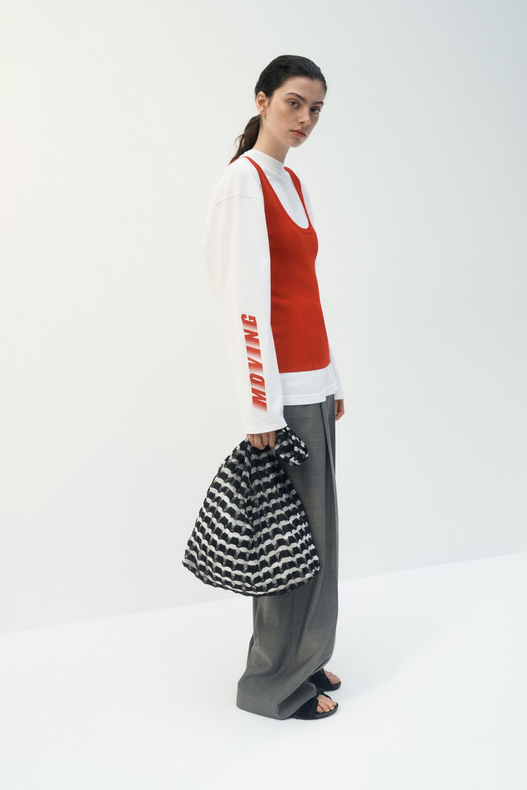 White jersey long-sleeve, red block lettering running down one cuff. Tomato red knit tank with a deep scooped neckline. Grey checked wide-leg trousers, pooling over the instep. Black-and-white crinkled tote with looped, wavy stripes. Flat black thong sandals. Walking back from a design fair with posters under one arm.