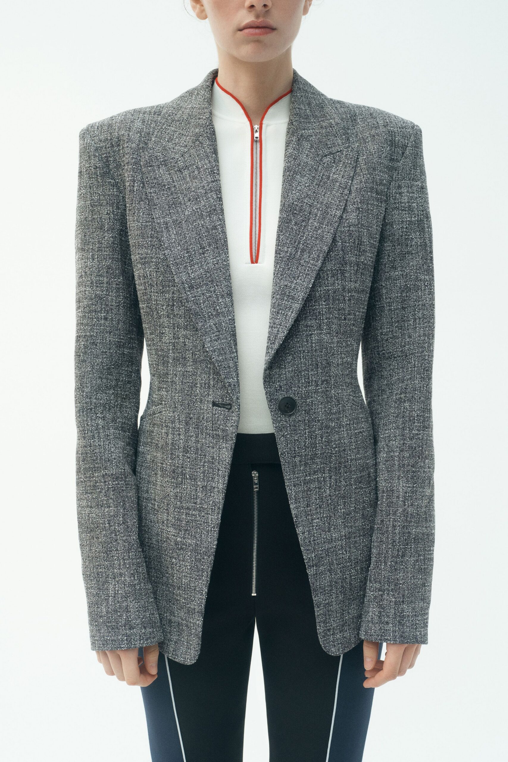 Grey salt-and-pepper tailored blazer with sharp padded shoulders and one dark button. White mock-neck zip top, red piping tracing the slit opening. Black slim trousers with a long silver fly zip and white side piping. For the first train out, station coffee burning through the paper cup.