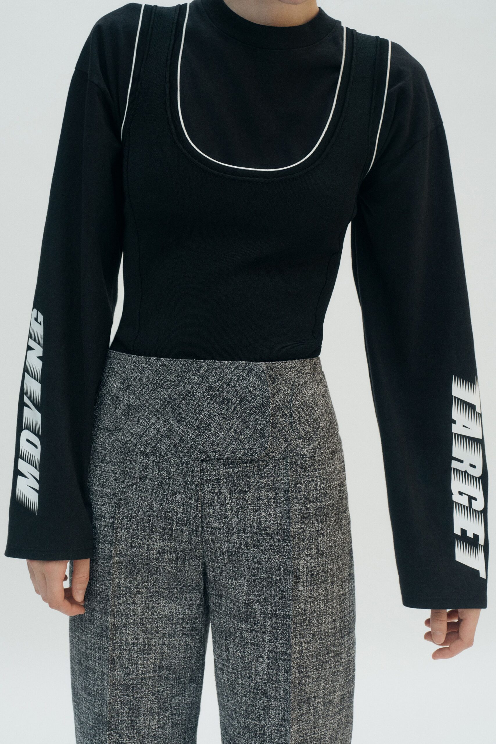 Black ribbed tank with white piping tracing the deep scoop. Black jersey tee, oversized sleeves stamped with blurred white type. Salt-and-pepper tailored trousers with a high rise and pressed front. Office lobby on Sunday, picking up the laptop you swore you'd leave there.