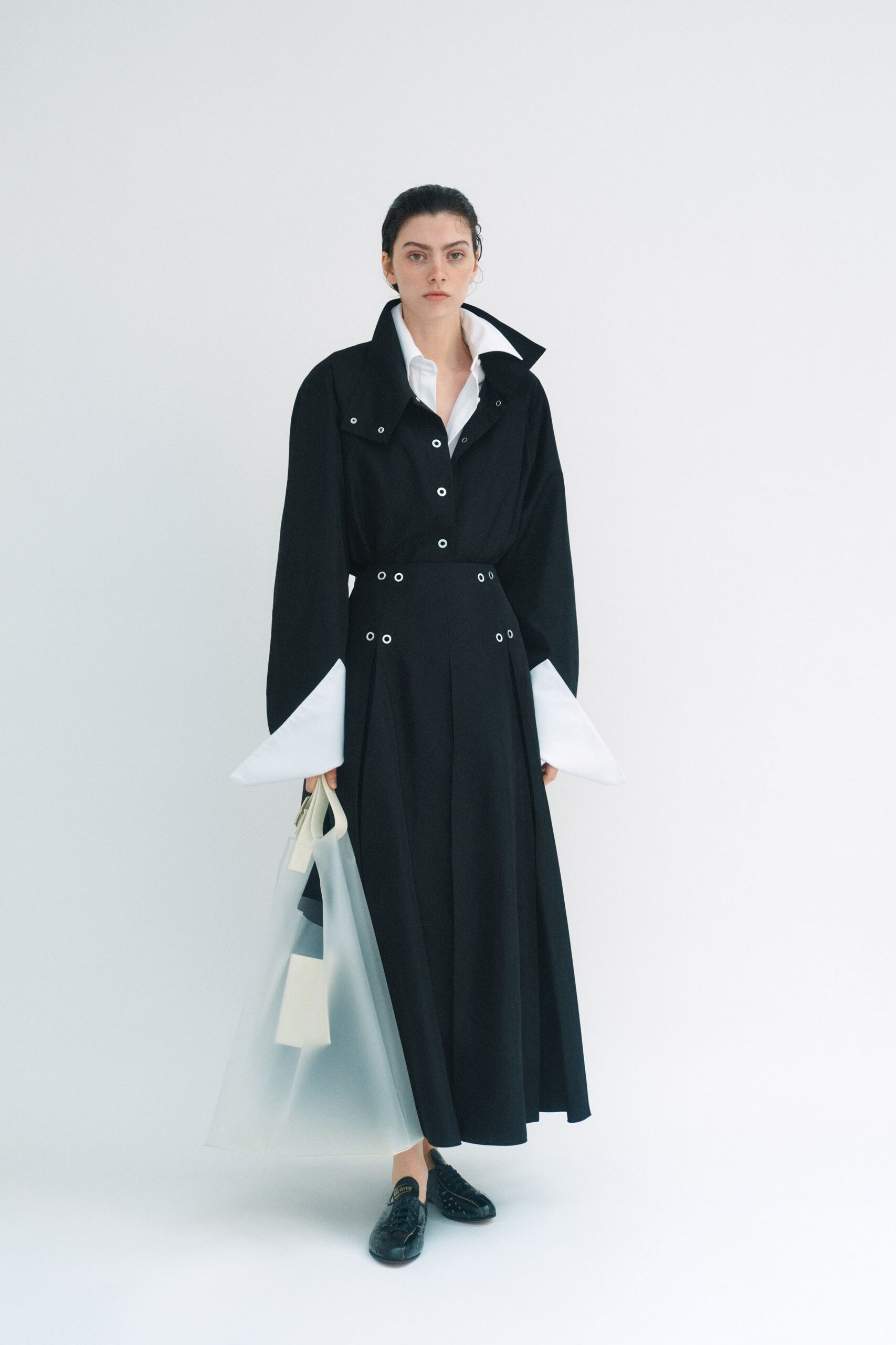 Black wool dress with a popped storm collar and silver eyelets. White poplin shirt, collar flared wide and cuffs jutting past the sleeves. Milky translucent tote with stark white handles. Black leather derbies, one toe nicked and creased. For carrying architectural plans across a lobby still smelling of fresh paint.