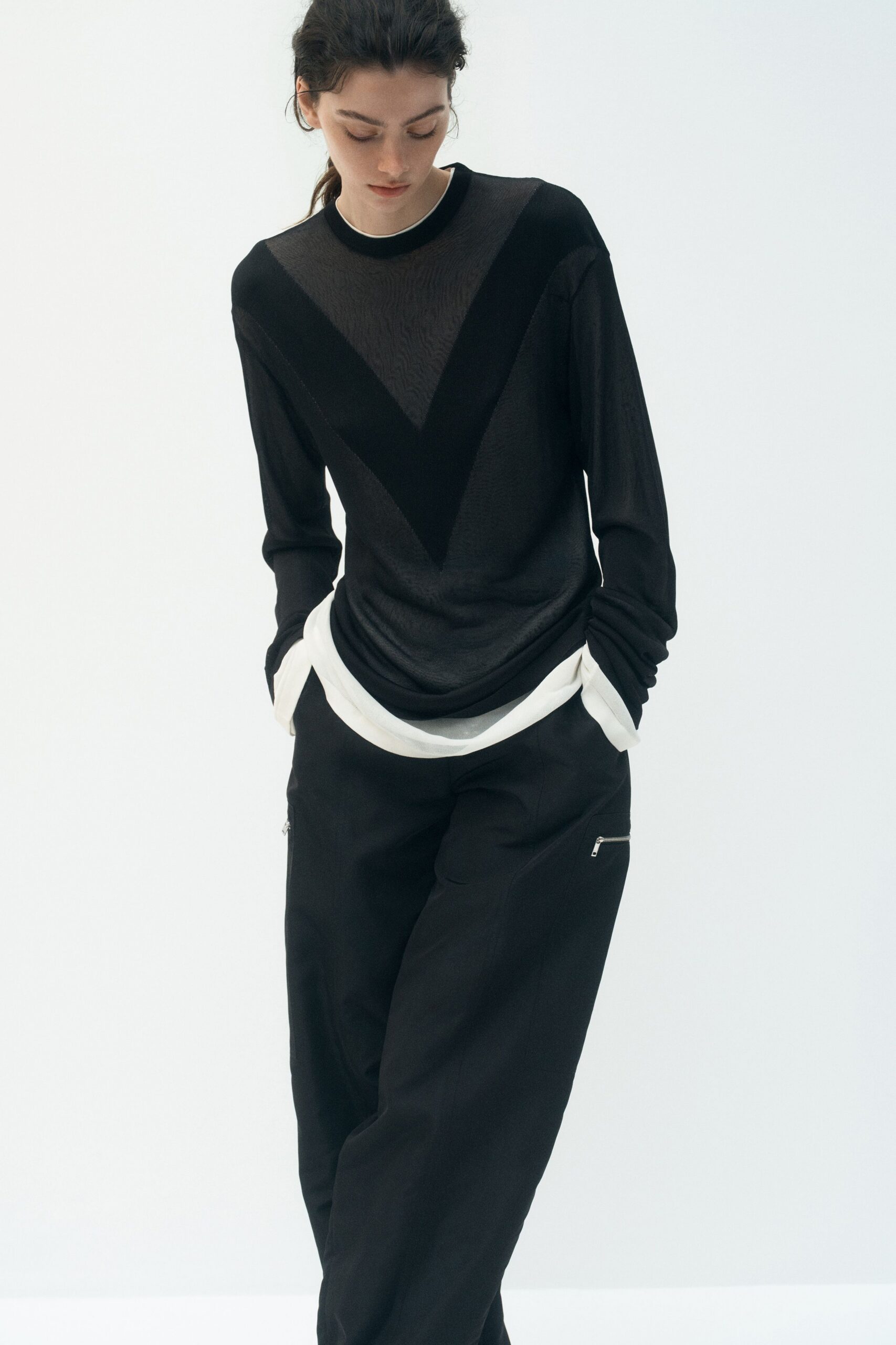 Sheer black knit top with a velvety V inset across the chest. White jersey hem and cuffs pushed out beneath it. Black straight trousers with small silver zips flicking at the thighs. First train Monday, coffee cooling between two gloved hands.